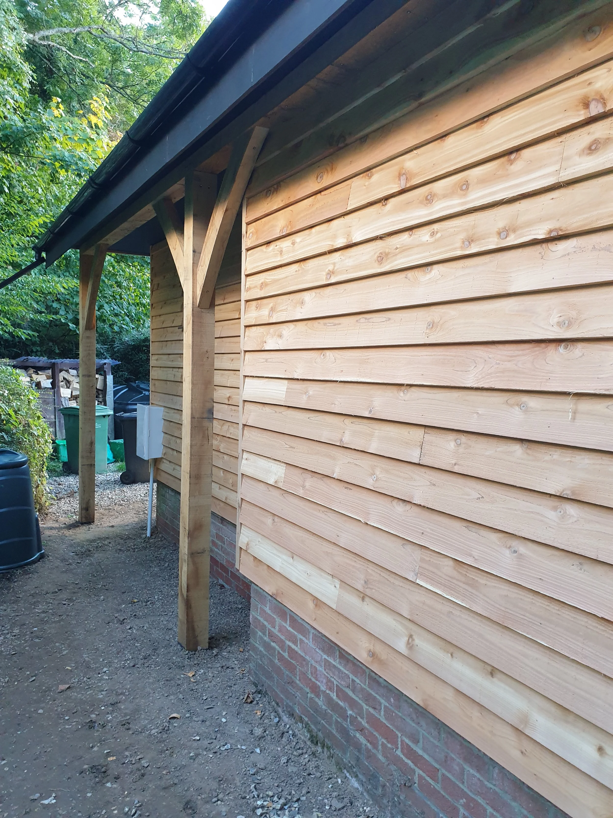 Side view of a house with fresh horizontal wooden siding and brick foundation, with a small overhang supported by wooden posts, surrounded by trees and garden tools.
