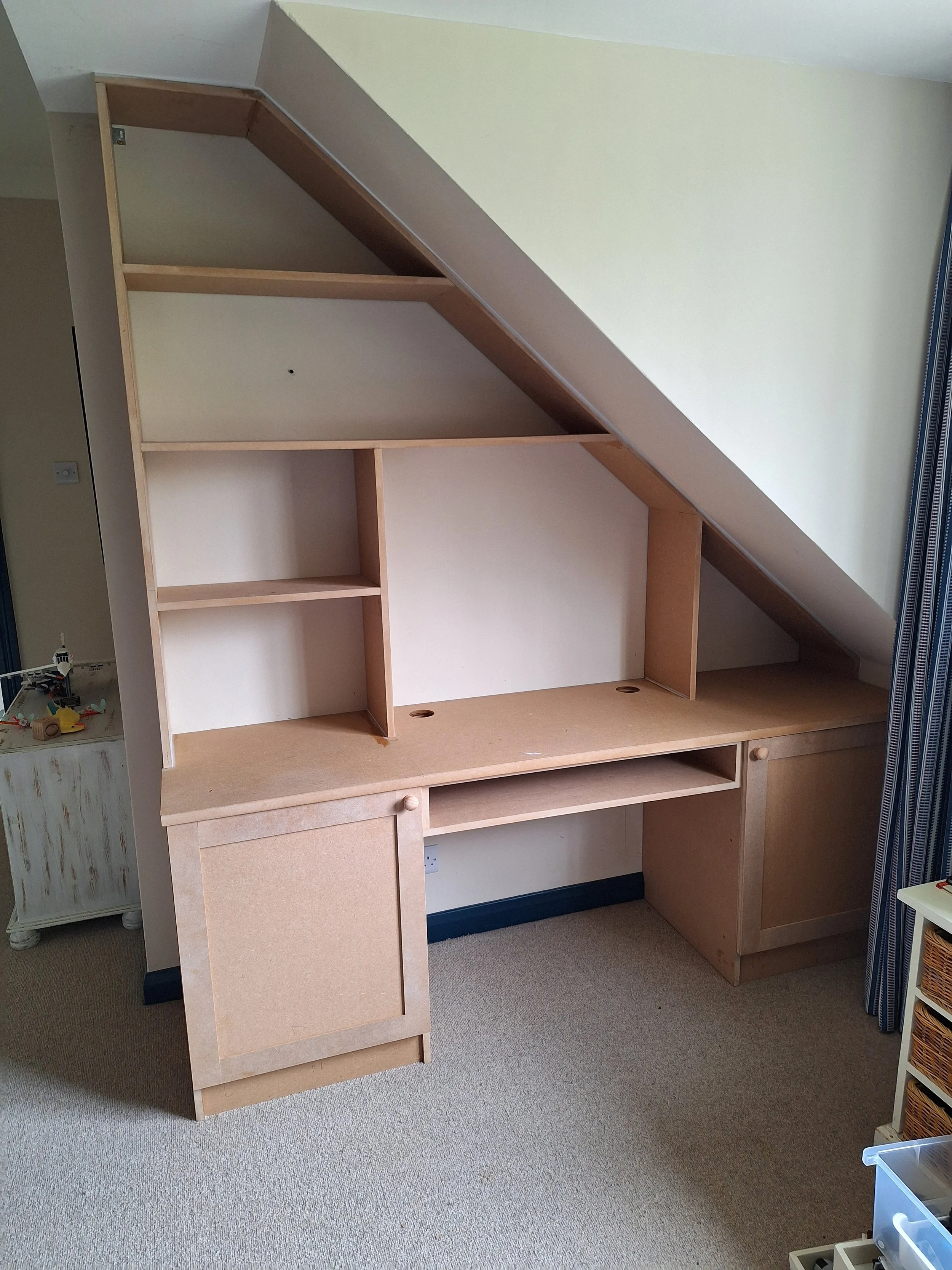 Unfinished custom-built desk or storage unit under a sloped ceiling in a room, made of unfinished wood with shelves and cabinets.