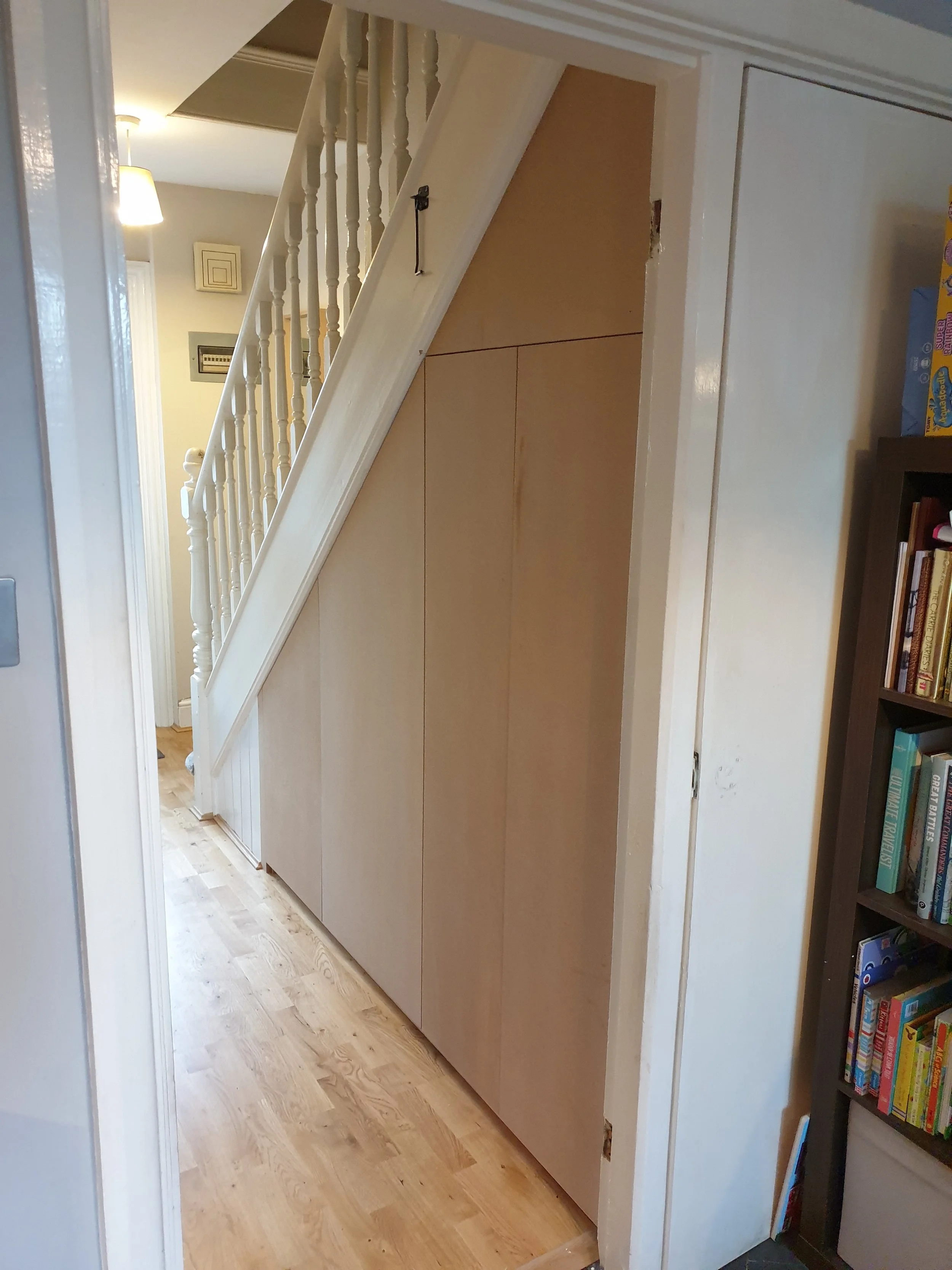 Interior view of a house showing a staircase with white spindles and a small hidden storage space beneath it, with a wooden door covering the storage area.