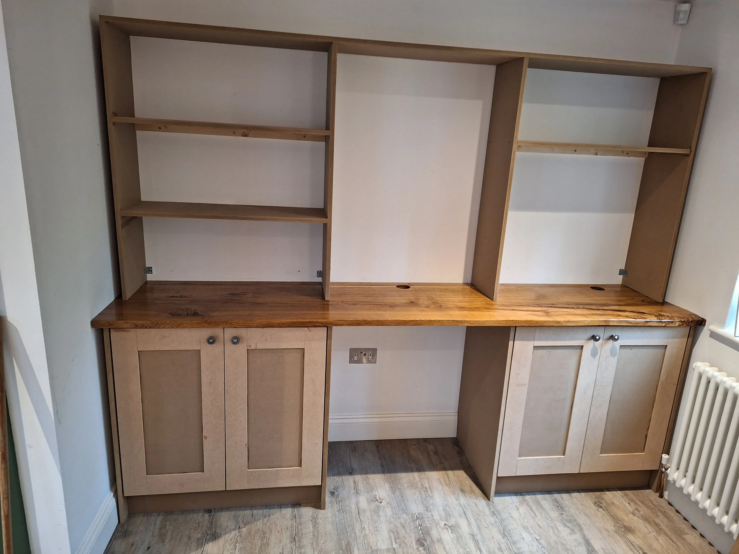 Wooden built-in desk with open shelving units above, two cabinet doors below, a power outlet, and a window nearby.