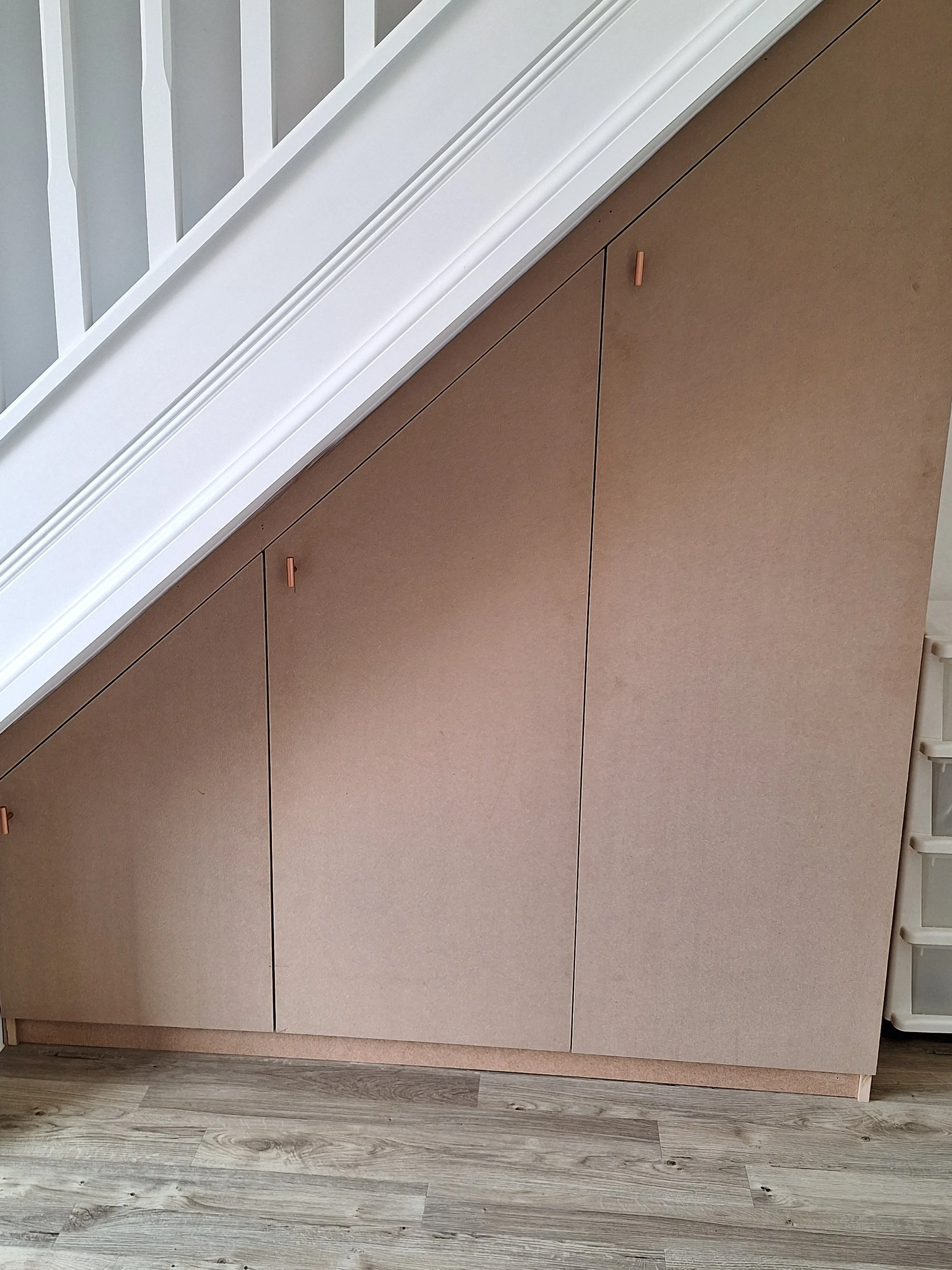 Brown cabinet beneath a staircase with a white railing, on a wooden floor.