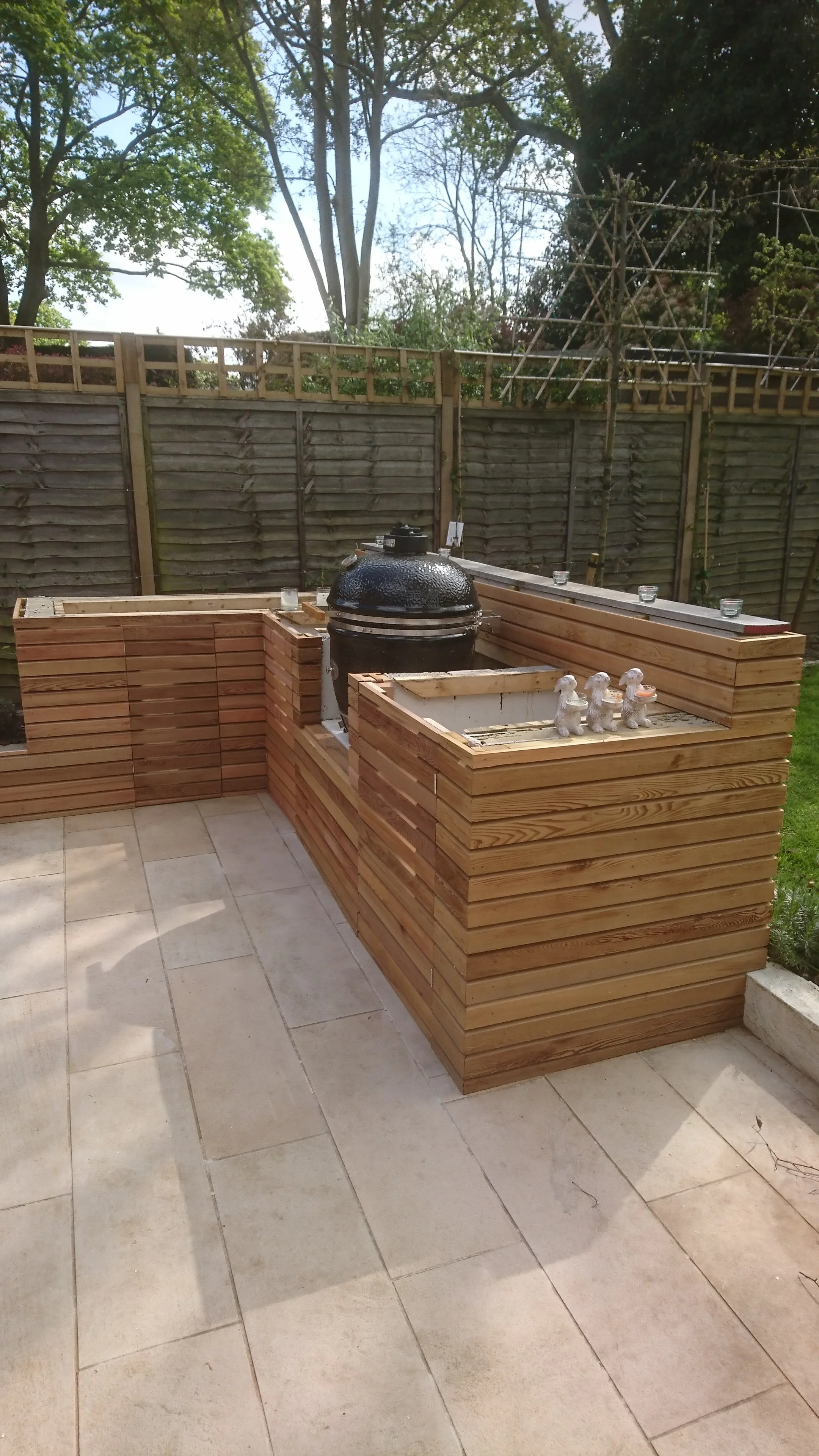 A backyard patio with a built-in wooden planter and a black grill, surrounded by a wooden fence and trees in the background.