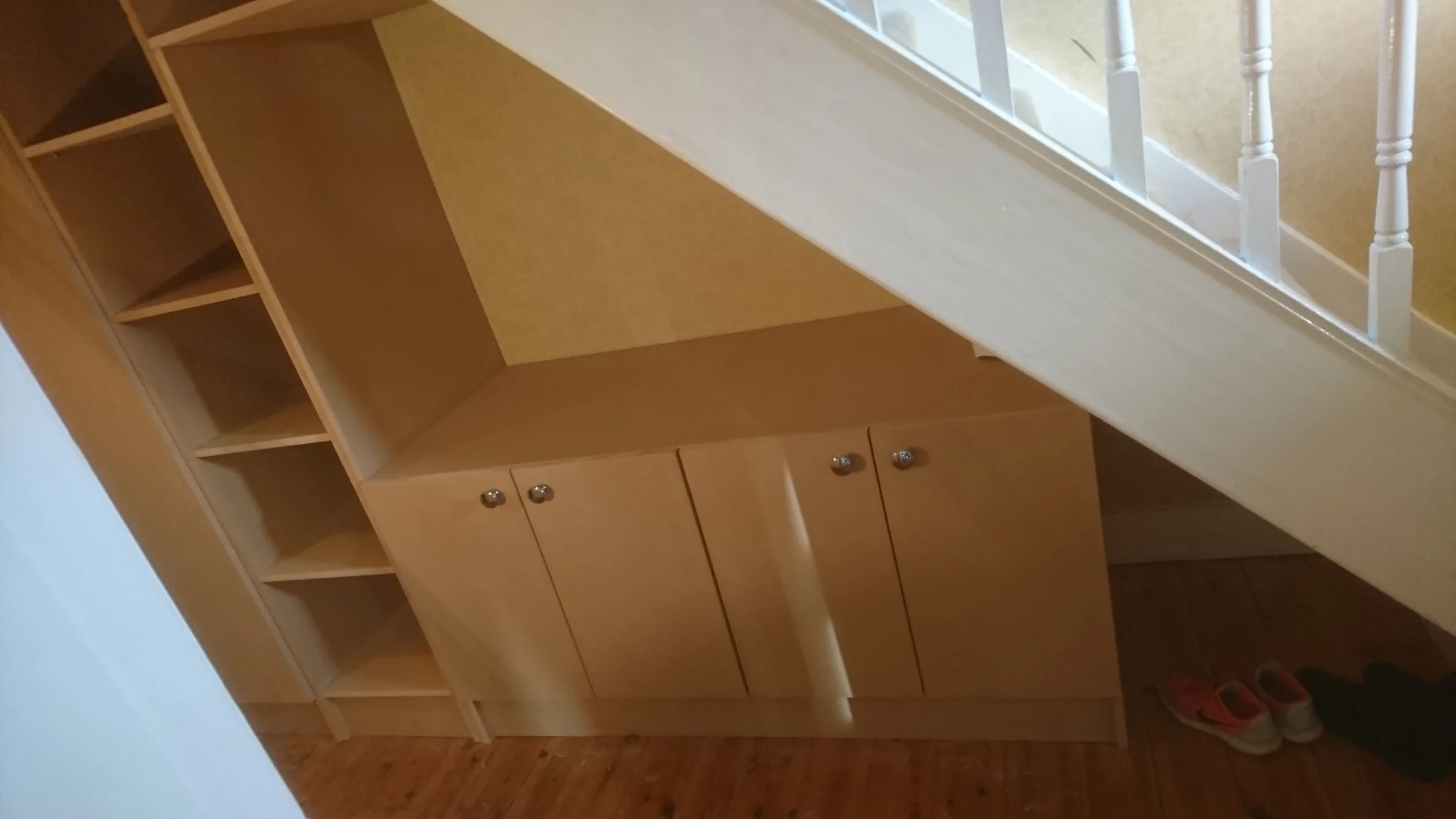 Wooden cabinet with shelves and closed storage under staircase, shoes on floor nearby.