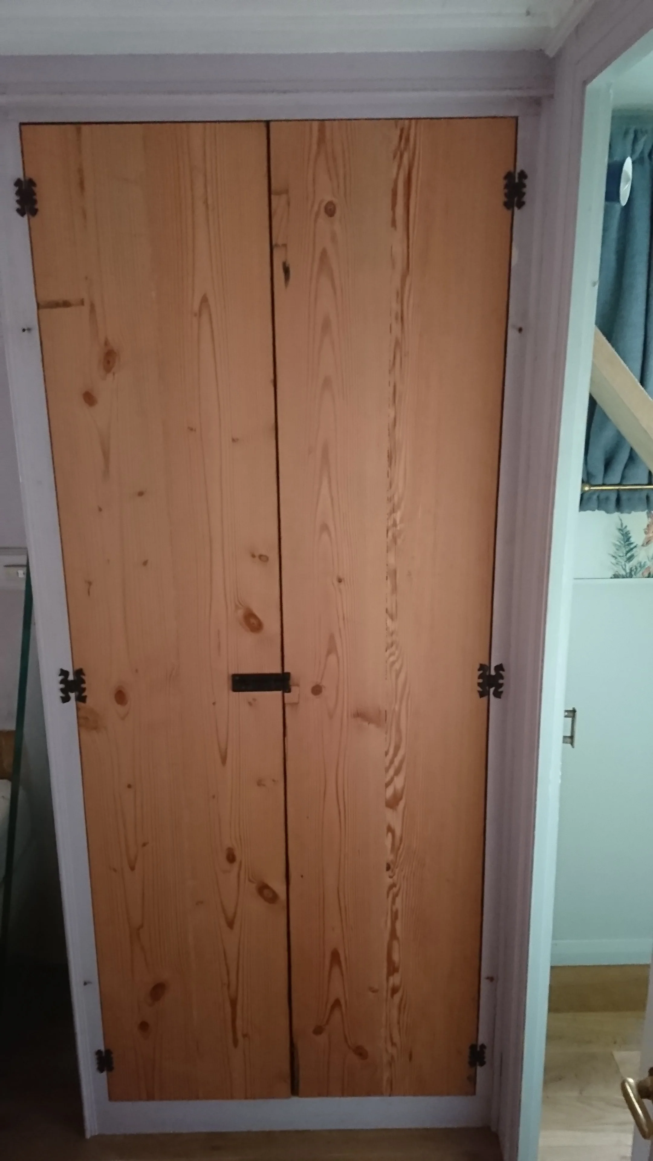 A wooden closet with two doors, featuring black hinges and a handle, situated within a white frame against a wall with white paneling.