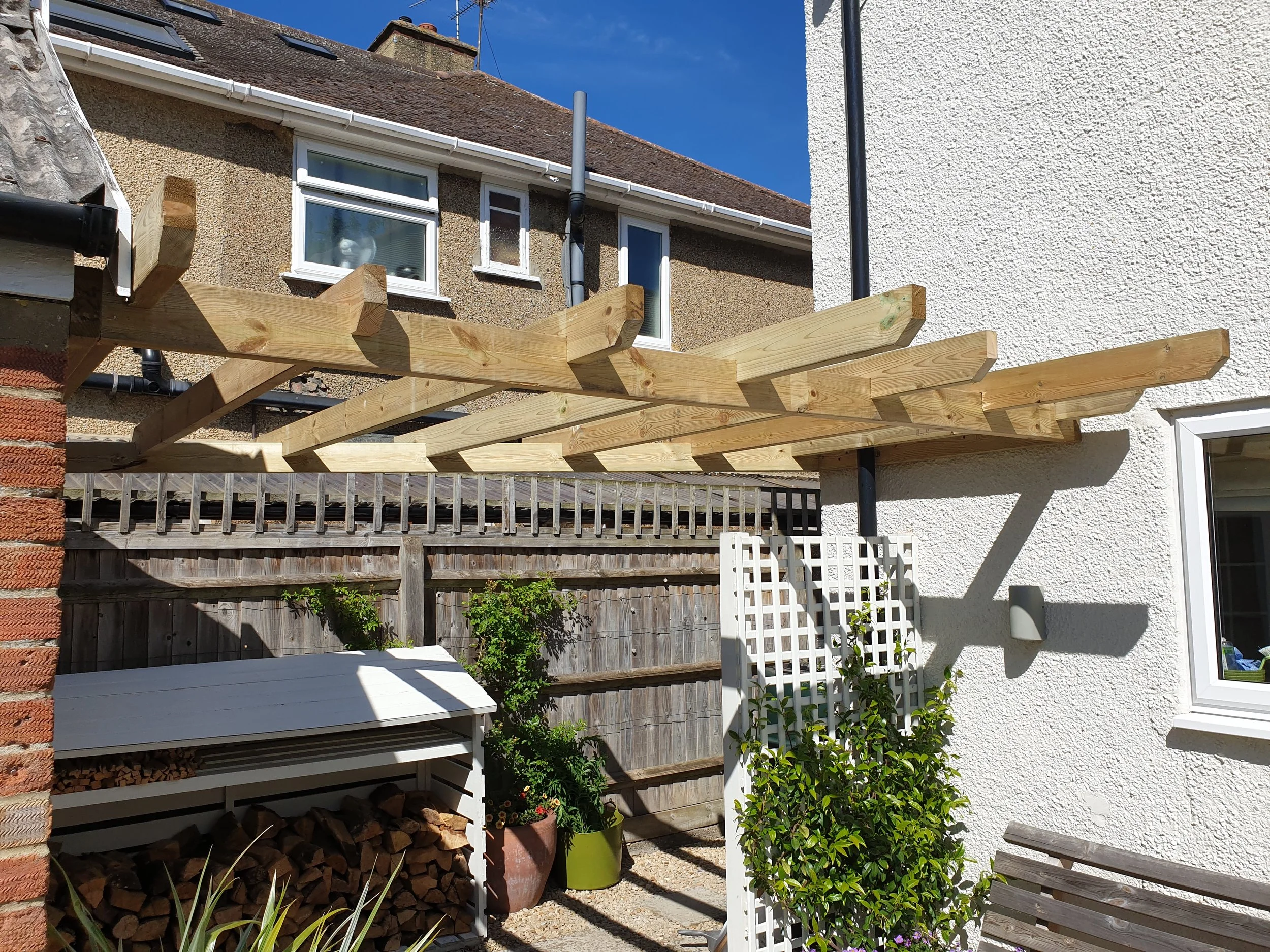 Unfinished wooden pergola structure attached to the side of a house in a backyard, with gardening plants and a woodpile underneath.