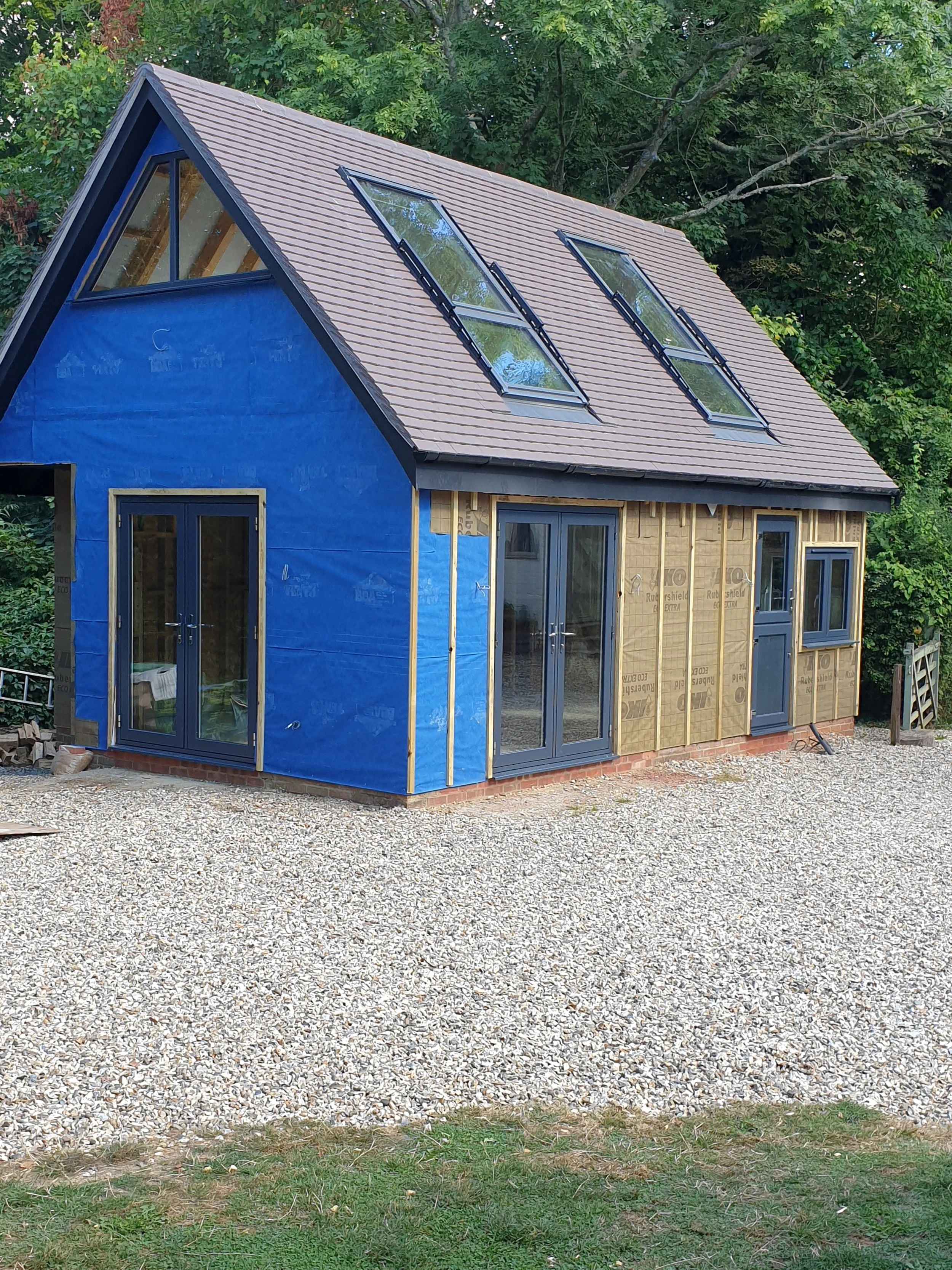 A house under construction with blue wall covering, multiple large windows with blue frames, and a gray tiled roof with skylights, surrounded by trees and gravel ground.