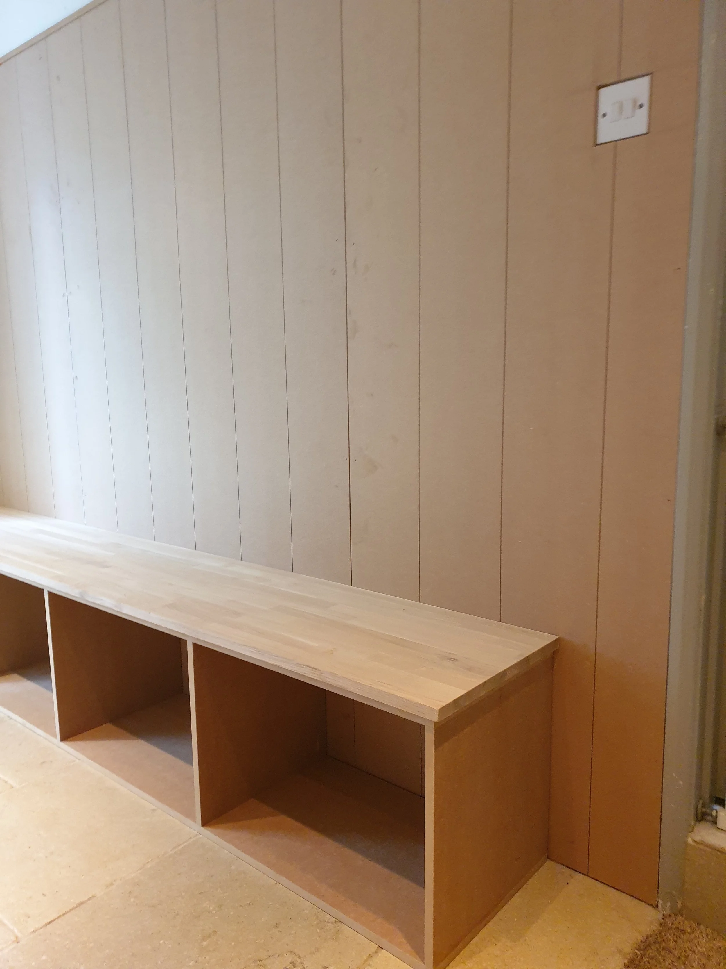 Wooden bench with open cubbies against a beige wooden wall with an electrical outlet.