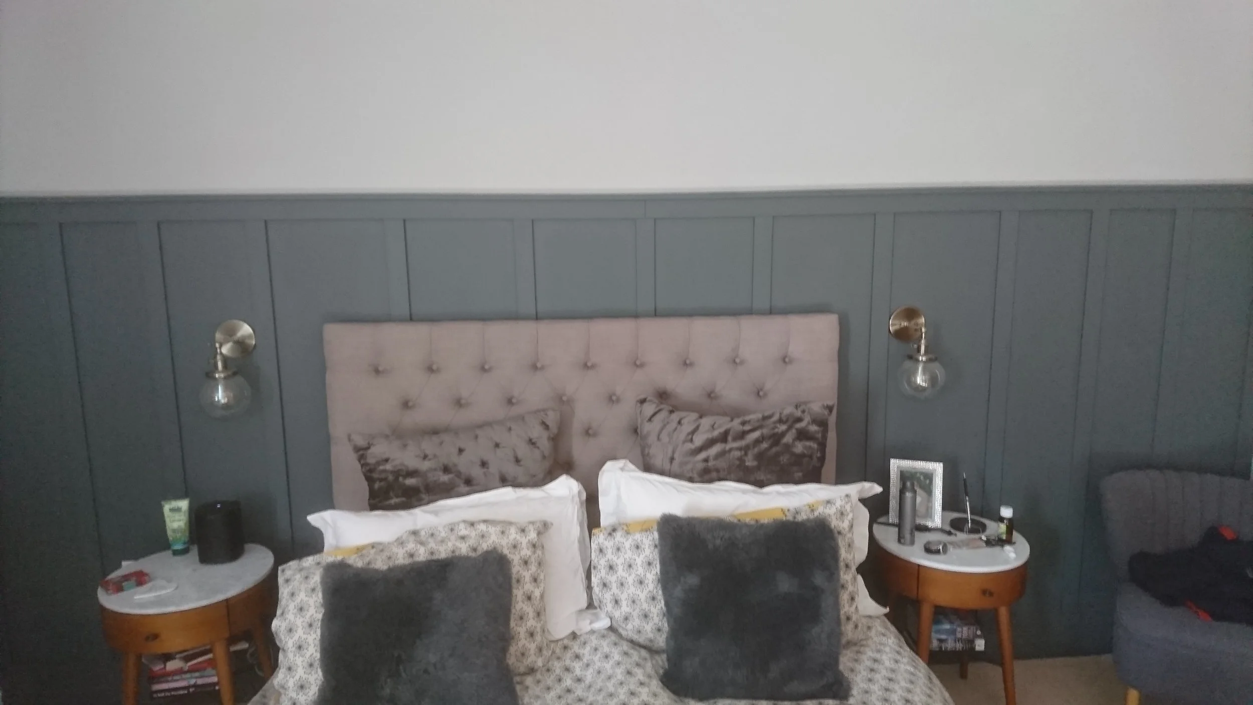 Bedroom with a bed, gray headboard, two dark gray pillows, patterned pillows, and two nightstands, one with a lamp, tissue box, and skincare products, and the other with a framed photo and more personal items, against a dark gray paneled wall.
