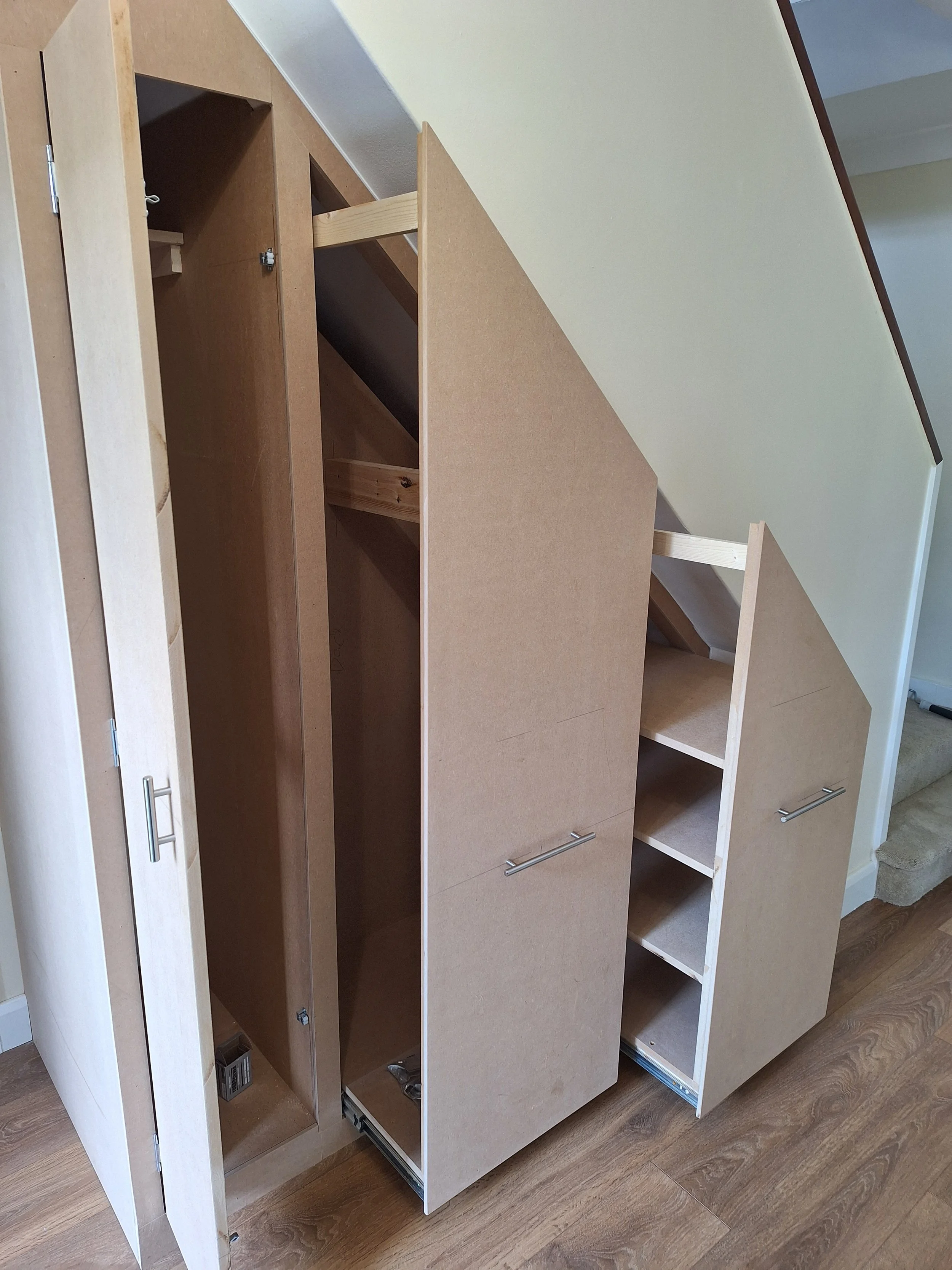 Unfinished custom-built closet or storage unit with sliding doors, shelves, and open compartments, under a slanted ceiling in a room with wood flooring.