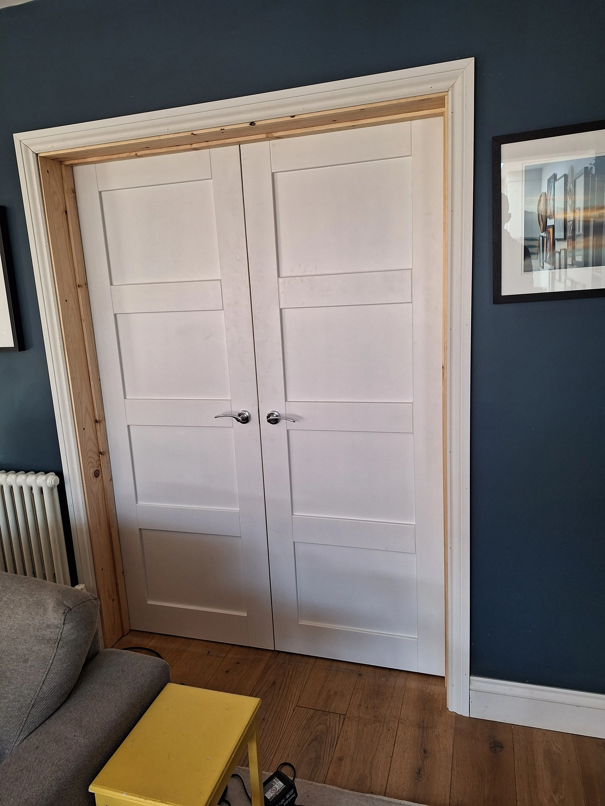White double doors with panel design, partially surrounded by natural wood framing, set against a dark blue wall in a room.