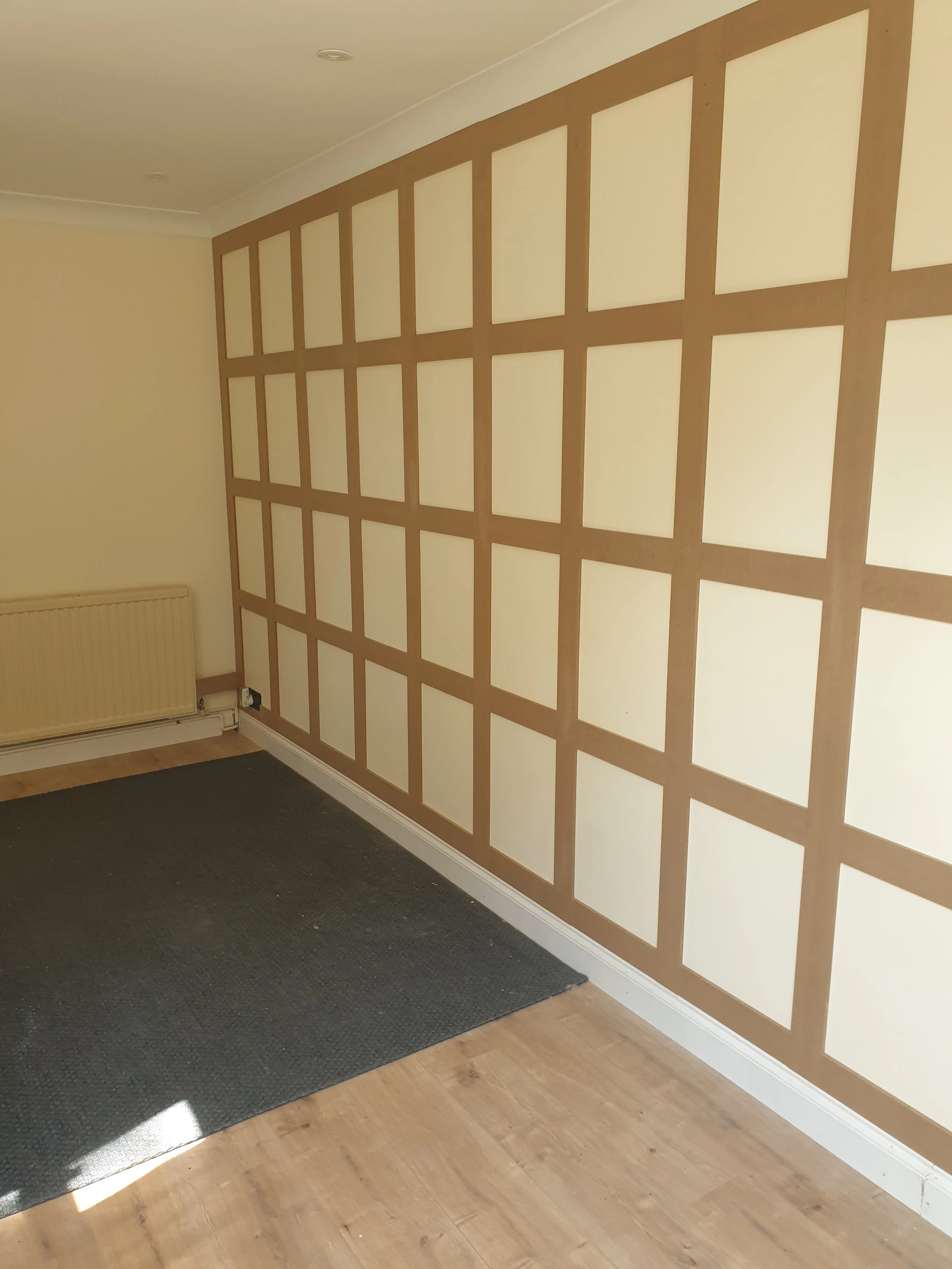 Interior view of a room with a wooden panel wall with a grid pattern, a radiator on the left, a dark rug on a light wooden floor, and a cream-colored wall.