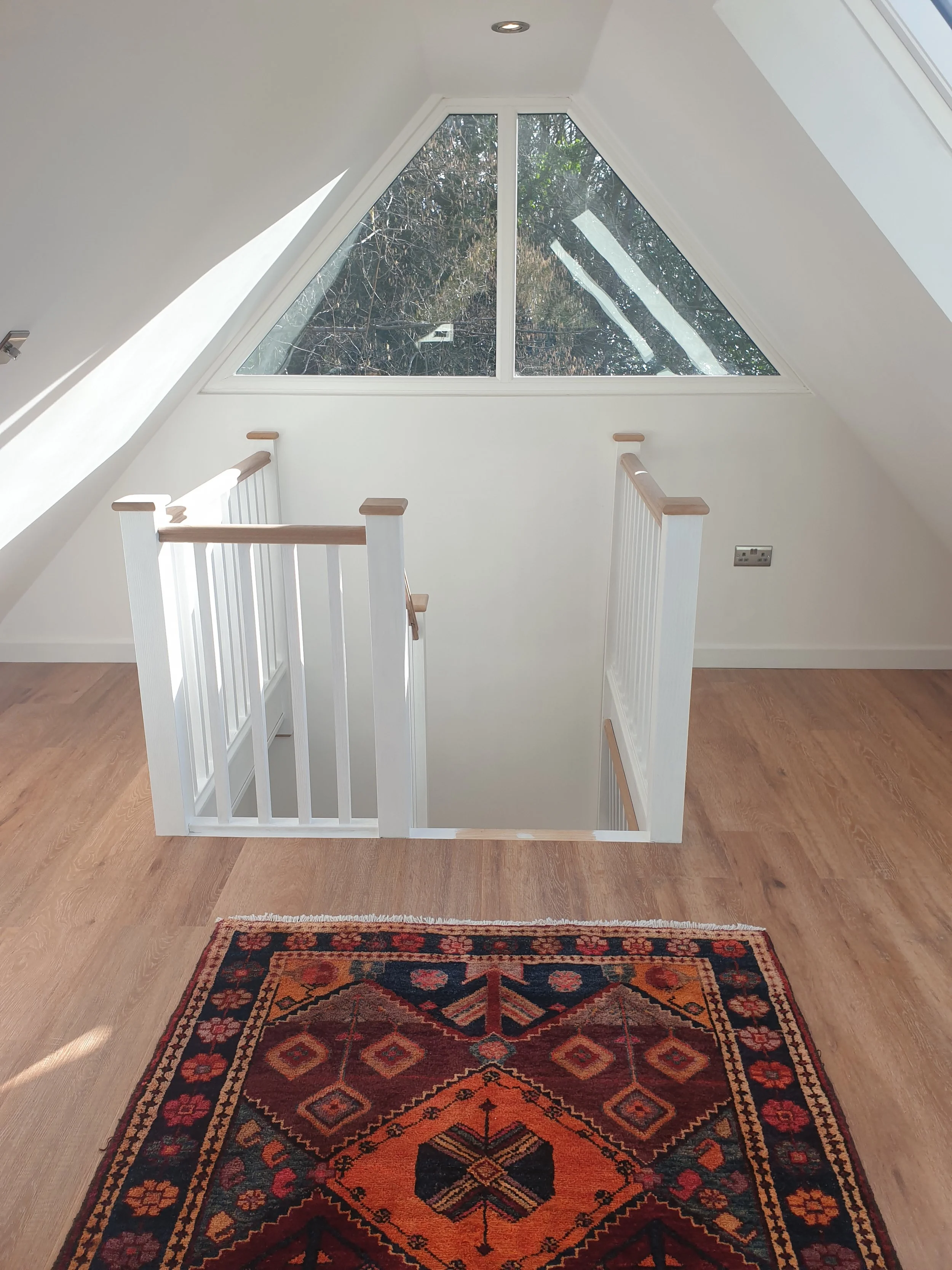 View of a staircase leading below, with a large triangular window above showing trees outside, inside a bright attic space with white walls, a colorful patterned rug, and hardwood flooring.
