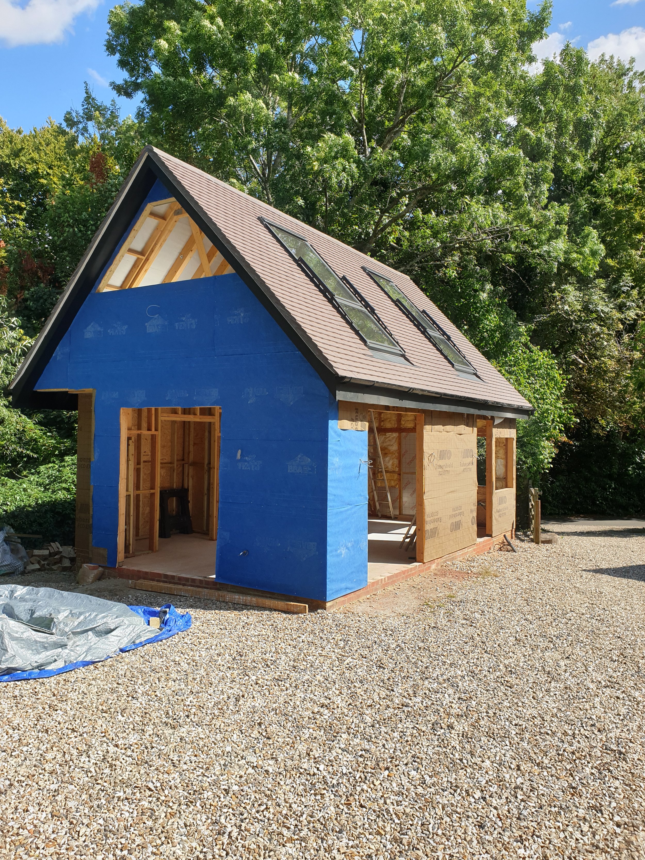 A house under construction with blue siding on one wall, exposed wood framing, a sloped roof with three skylights, and surrounded by trees on a gravel lot.