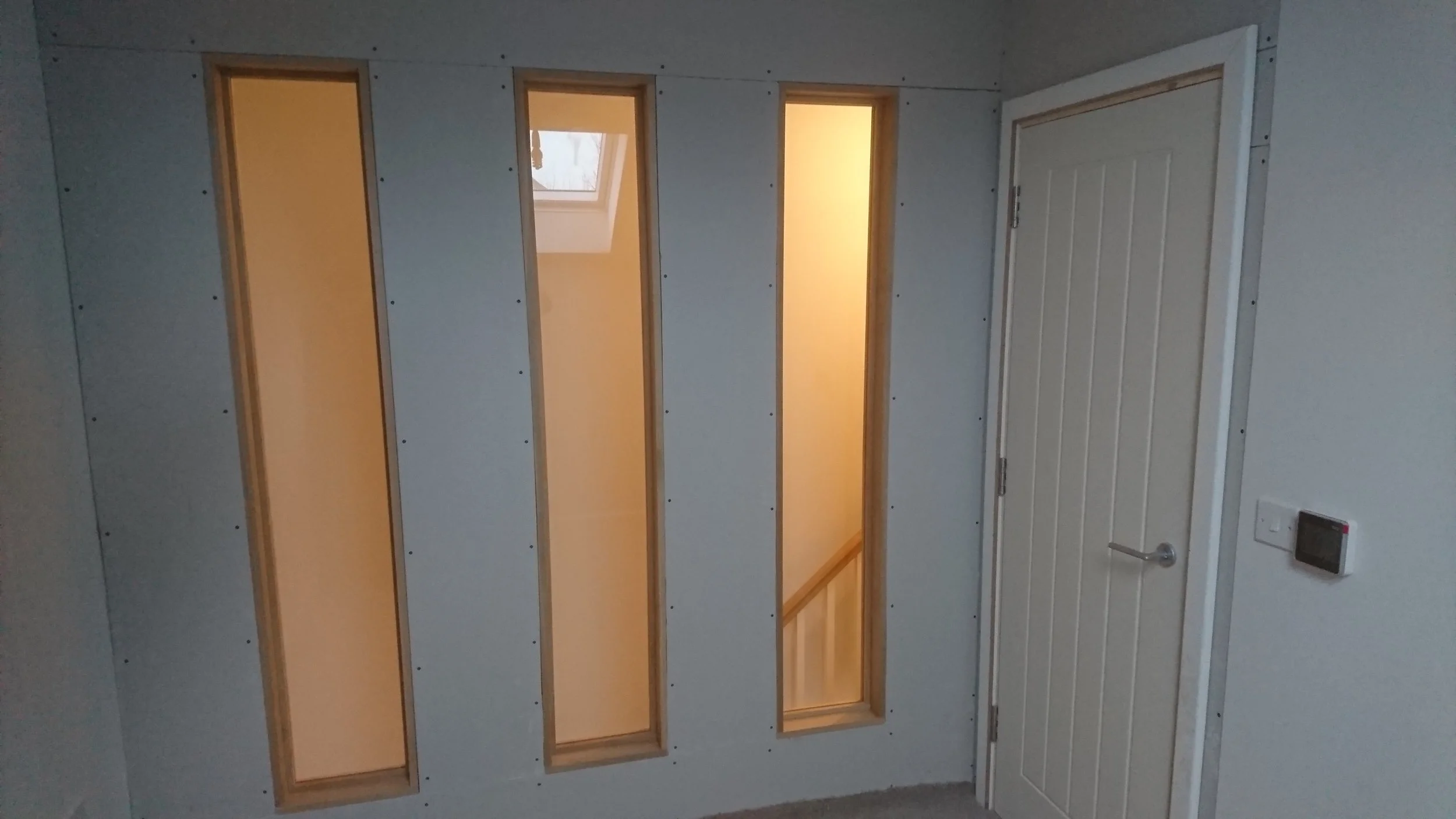 Interior view of a partly constructed room with three tall, narrow windows and a closed white door, with drywall panels on the walls and a thermostat on the right wall.