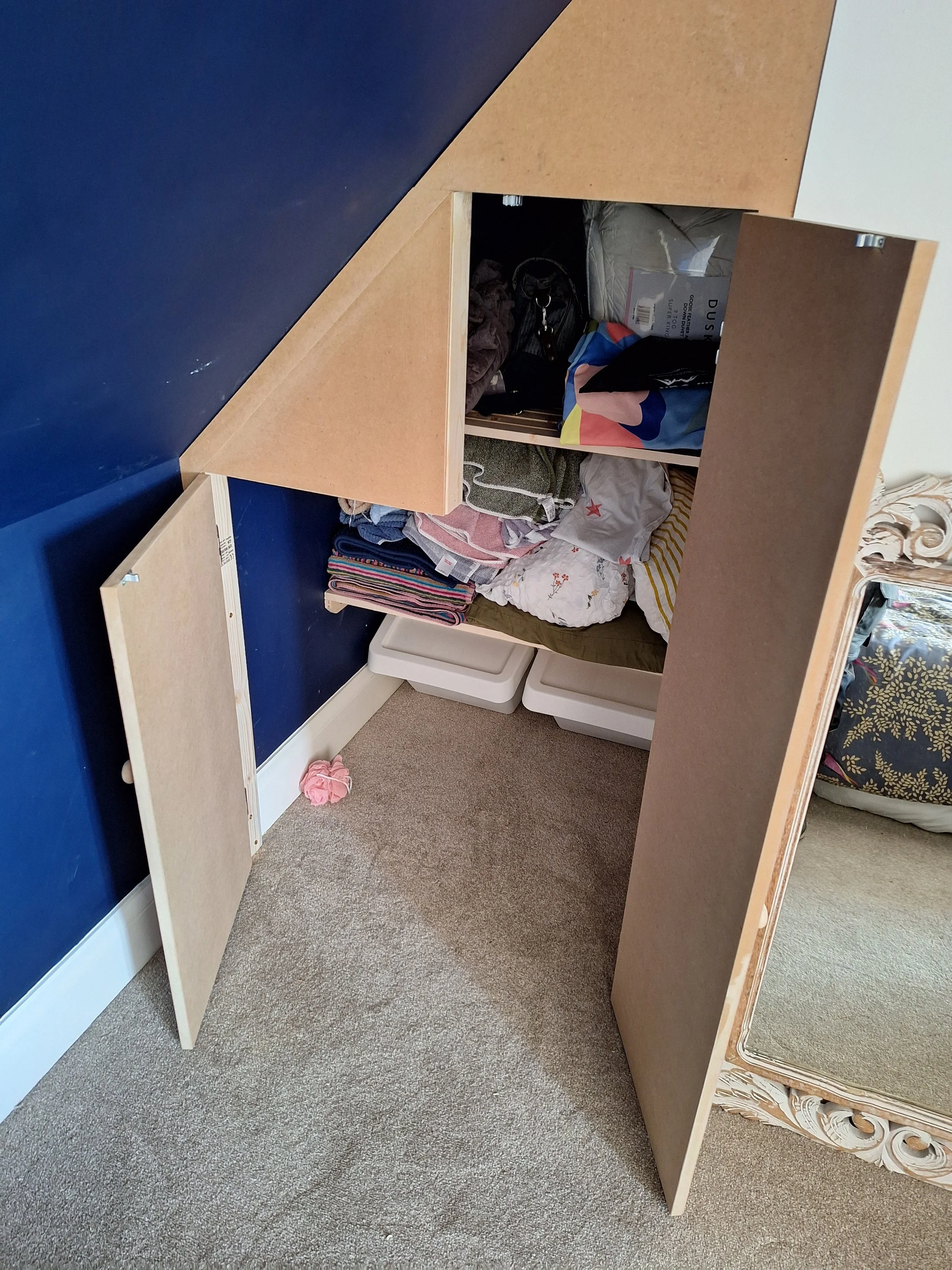 The image shows a small, partially assembled wooden cabinet or shelf unit with its doors open, revealing various folded clothes, bags, and other miscellaneous items inside. The piece of furniture is placed on a beige carpet near a large ornate mirror