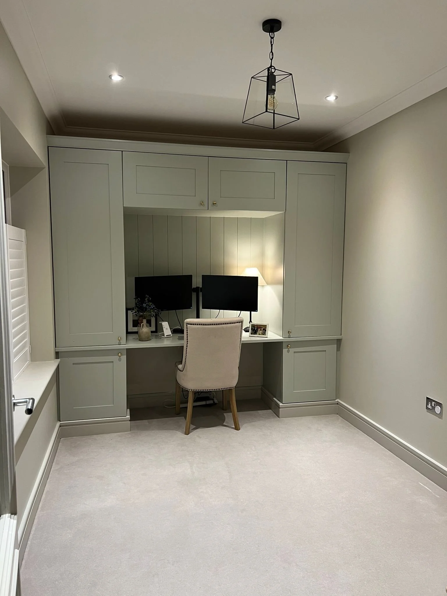 A home office with built-in shelves, a desk with dual monitors, a beige upholstered chair, a lamp, framed photos, and neutral-colored walls and carpet.