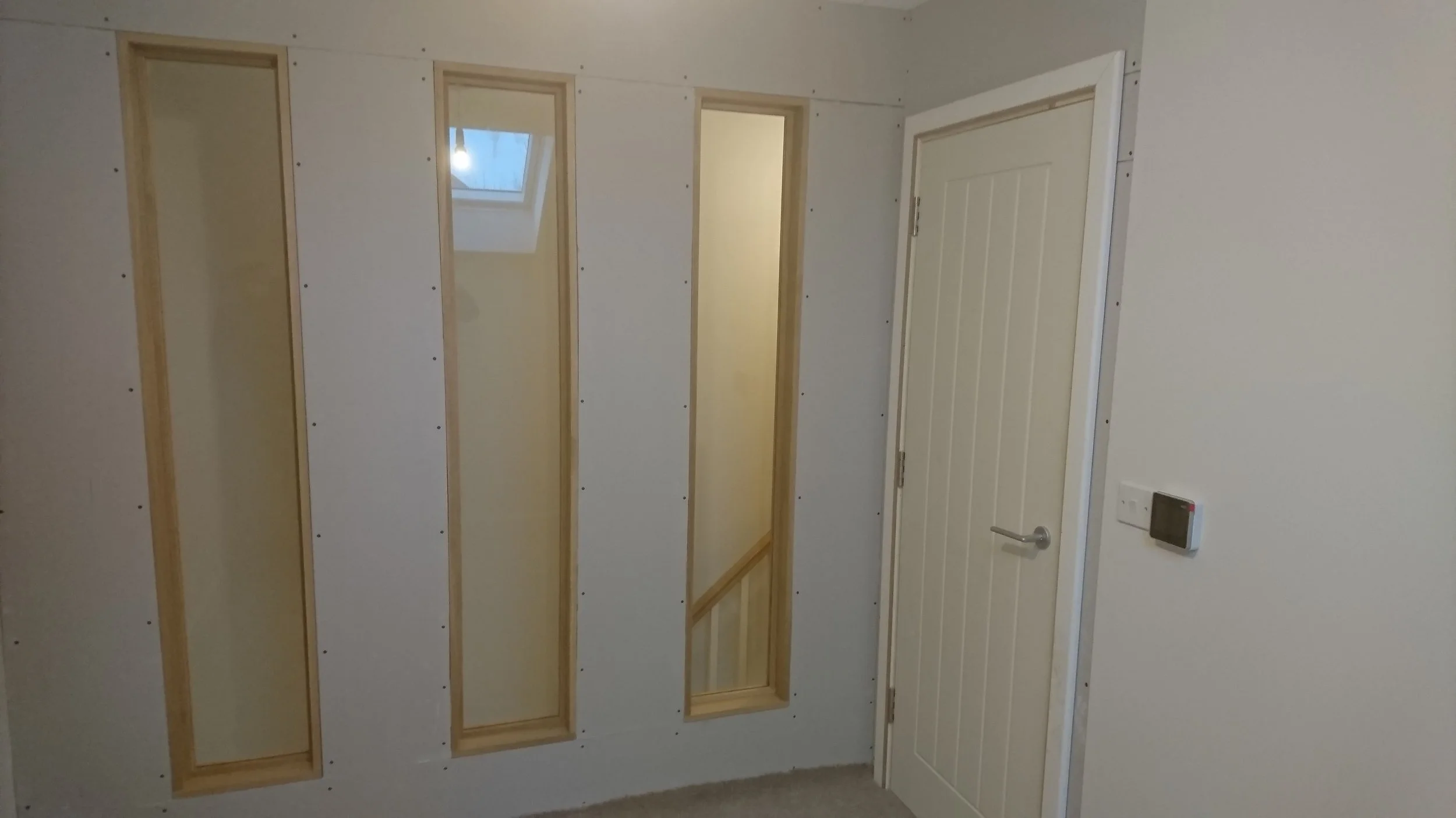 Interior view of a room with partially installed wall panels, four vertical rectangular cutouts framed in wood, a white door, and a wall-mounted thermostat.