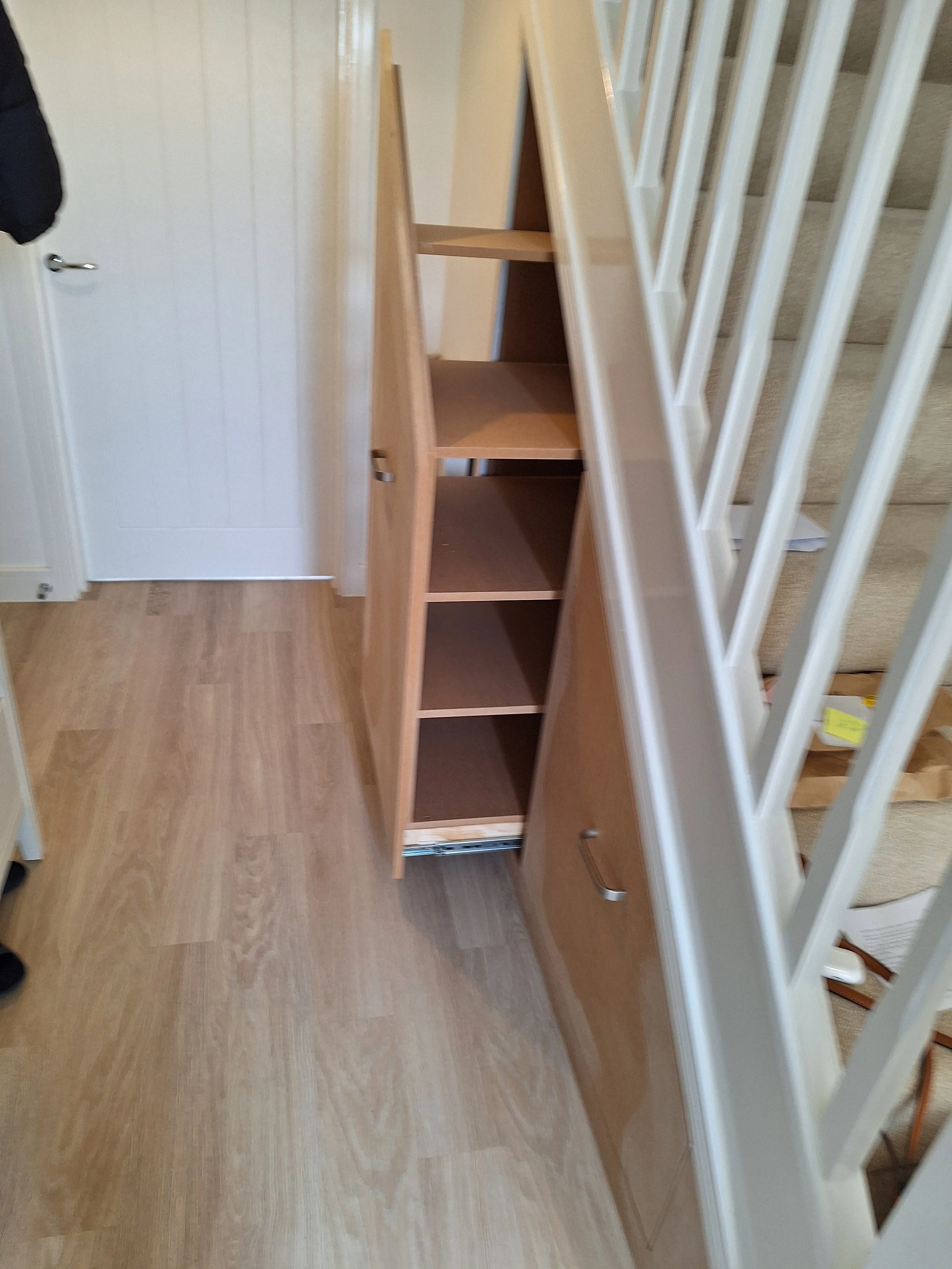 Empty wooden bookshelf with four shelves, located near a staircase with white railing, in a room with light-colored wood flooring and a white door.