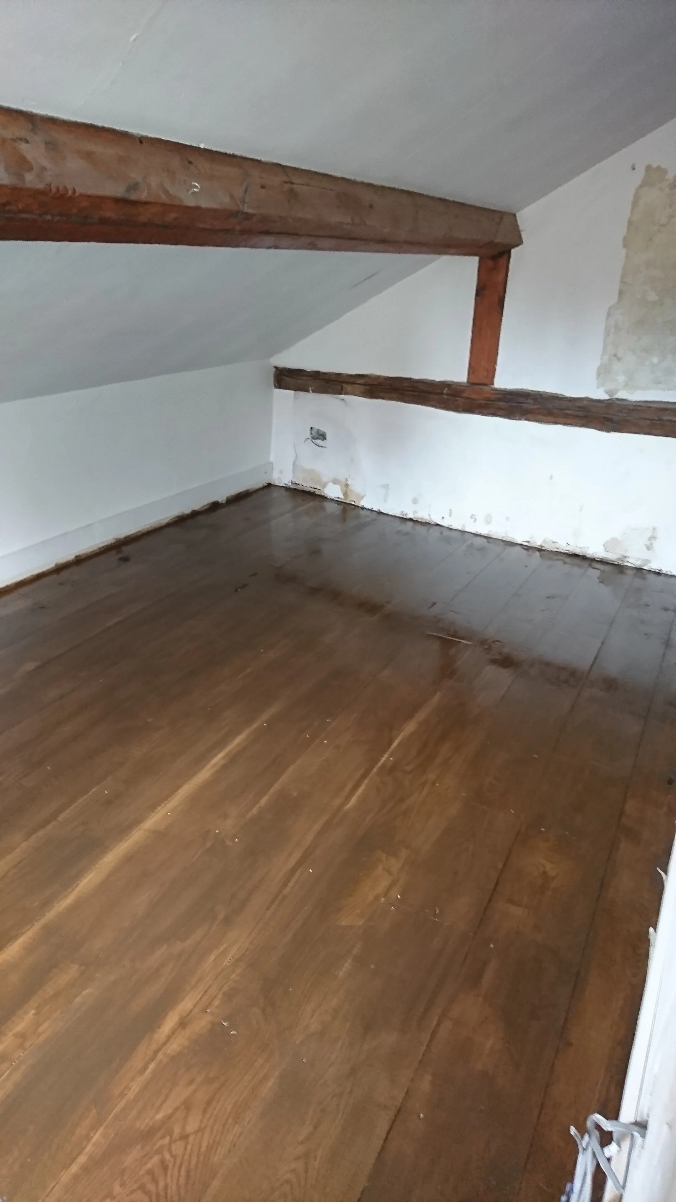 Room with sloped ceiling, exposed wooden beams, and a hardwood floor. There is a wall with peeling paint and an electrical outlet.