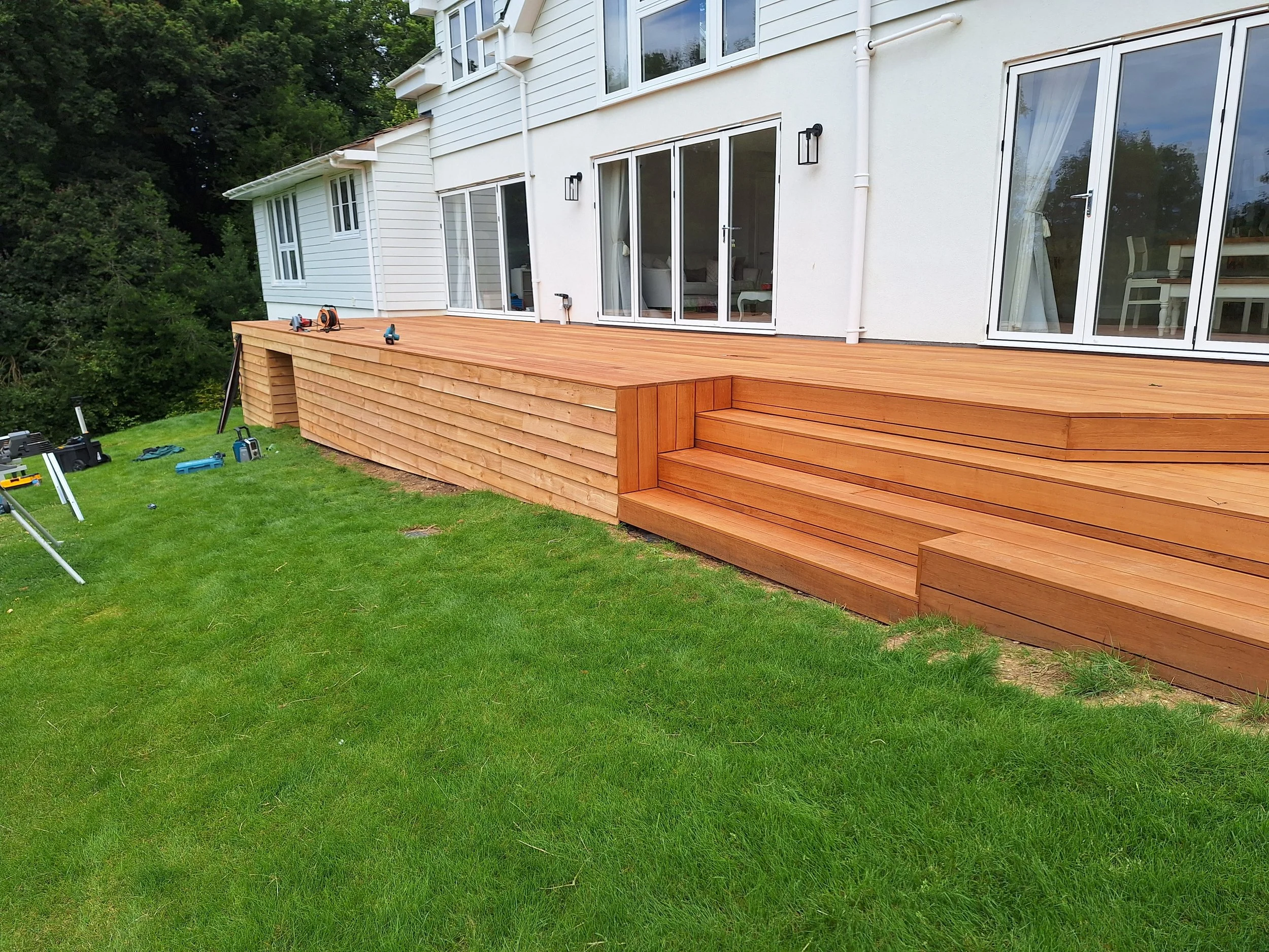 Newly constructed wooden deck with steps attached to a white house with sliding glass doors, situated in a green backyard.