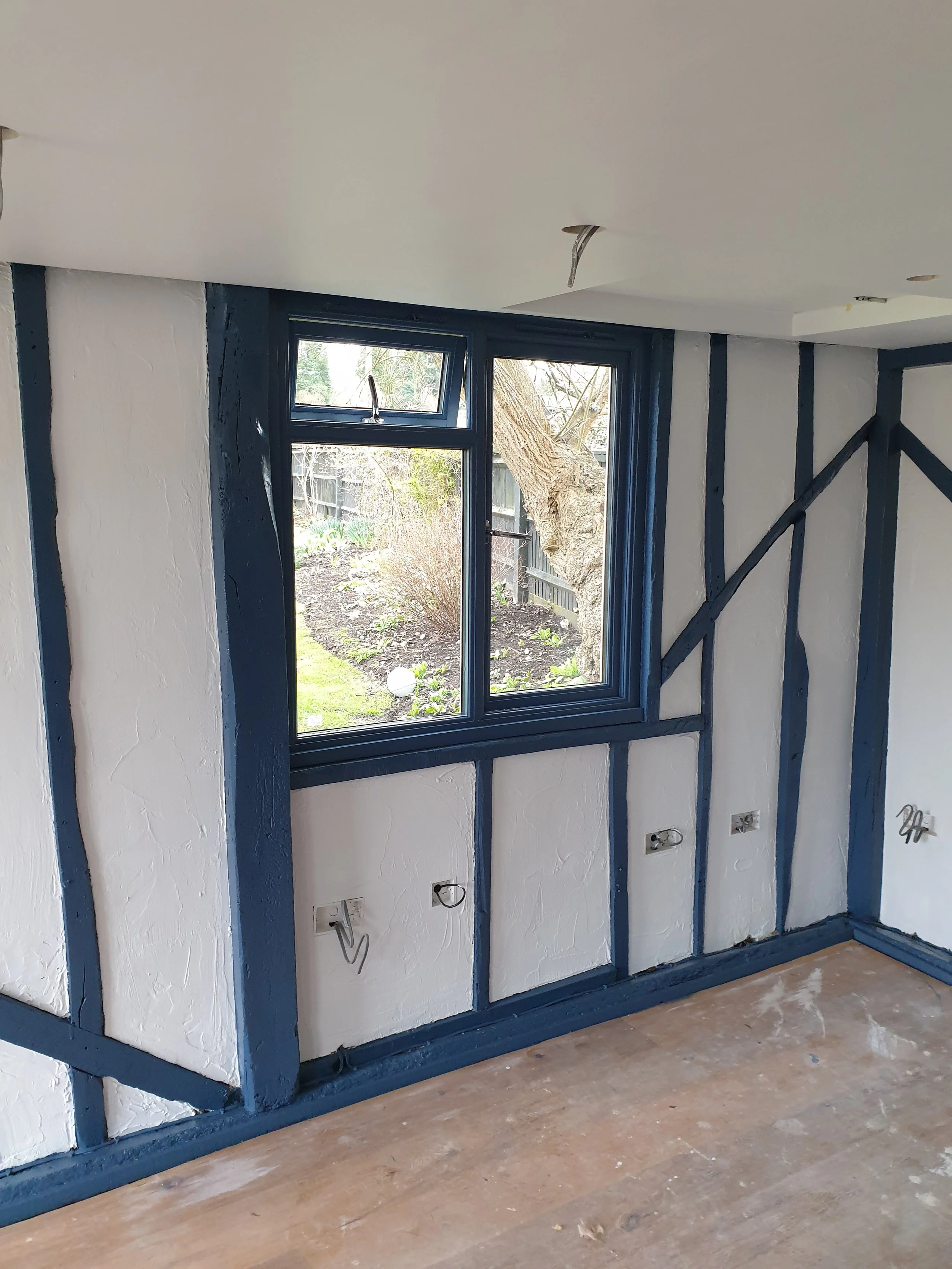 Interior of a room under renovation with a large window, blue painted beams, and electrical outlets, with a view of a garden outside.