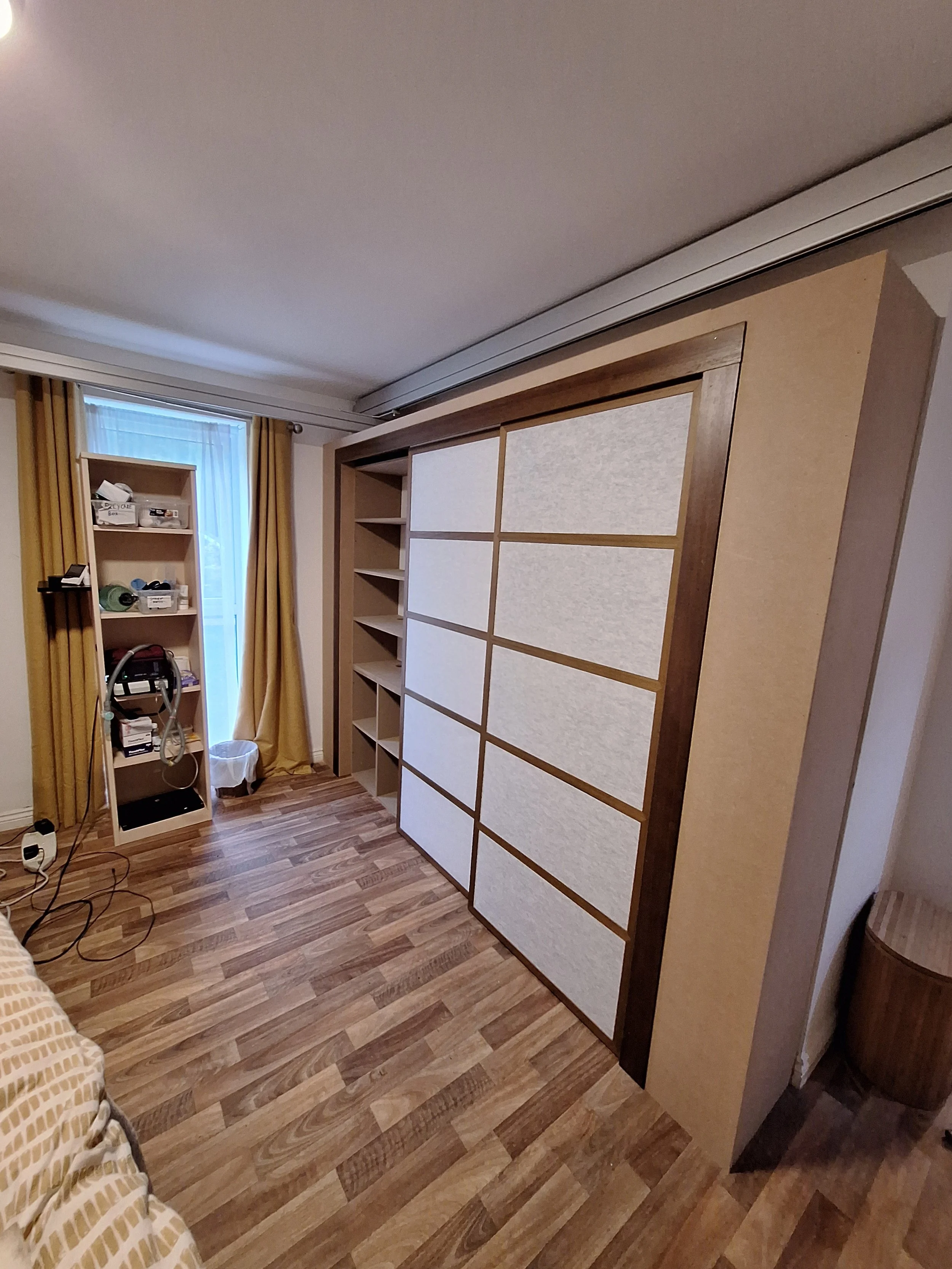 Room with wooden flooring, a wardrobe with sliding doors, a shelving unit, and a window with mustard curtains.