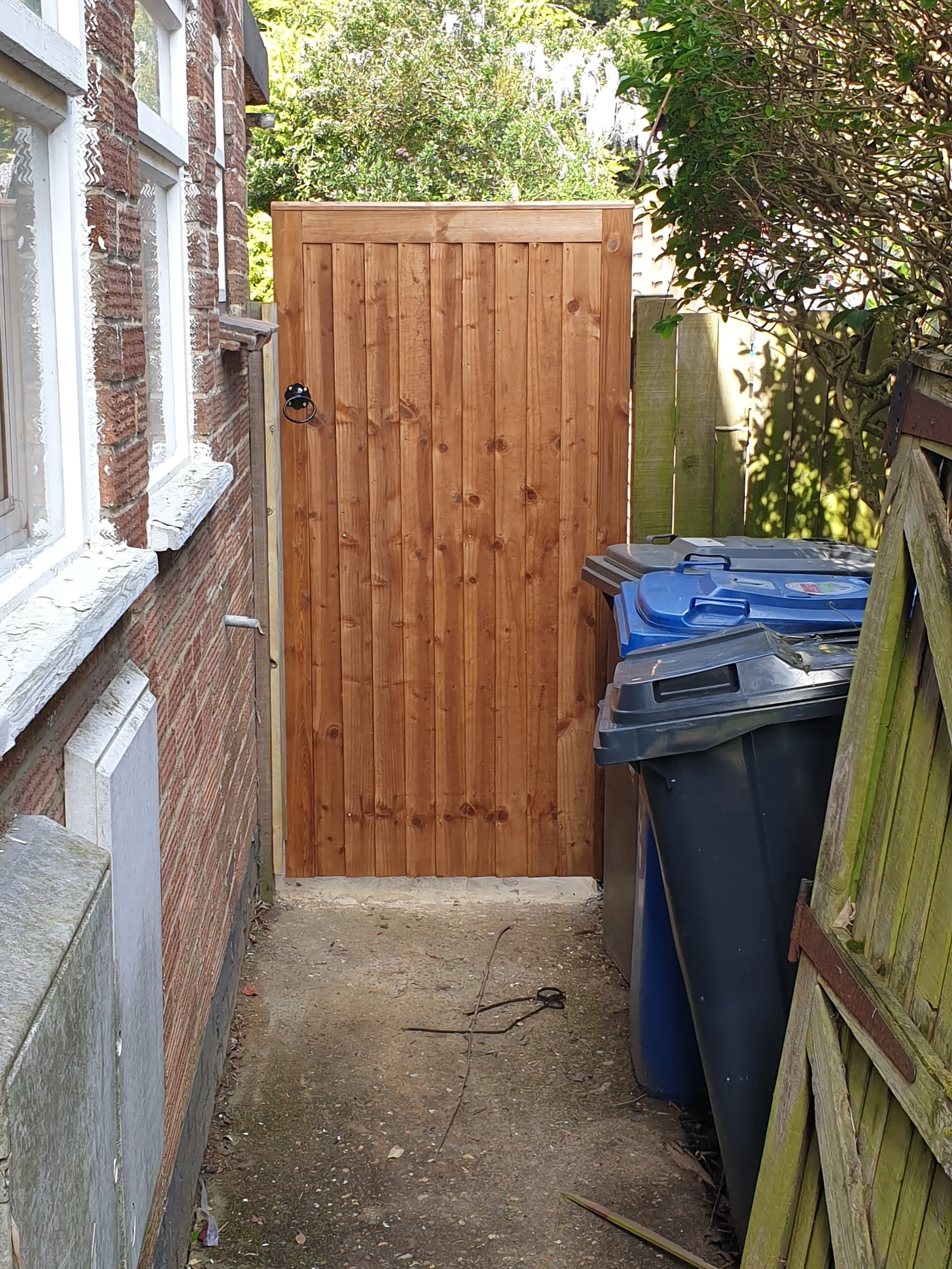 A small outdoor area with a wooden gate at the end, flanked by a brick wall on the left and a weathered wooden fence on the right. There are three trash bins with blue and black lids in front of the fence. Green leafy trees are visible behind the gat