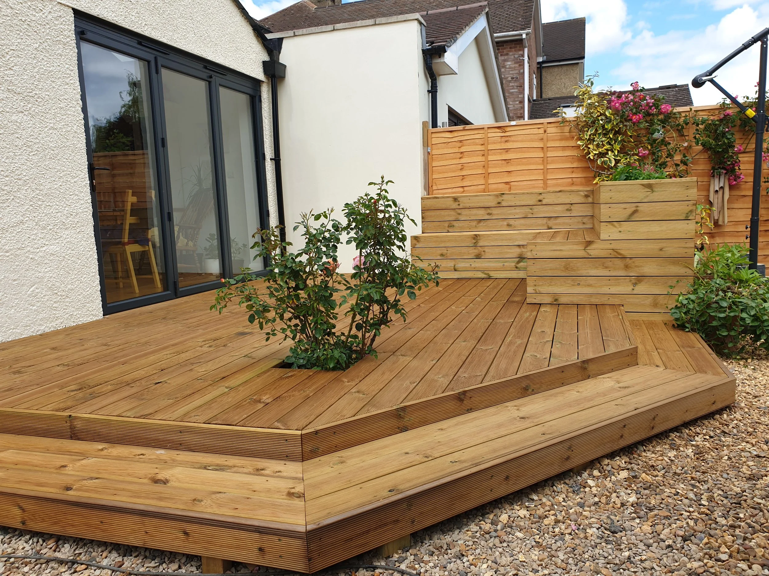 A newly built wooden deck with raised planter beds and small plants, adjacent to a house with large glass sliding doors, enclosed by a wooden fence with flowering plants.