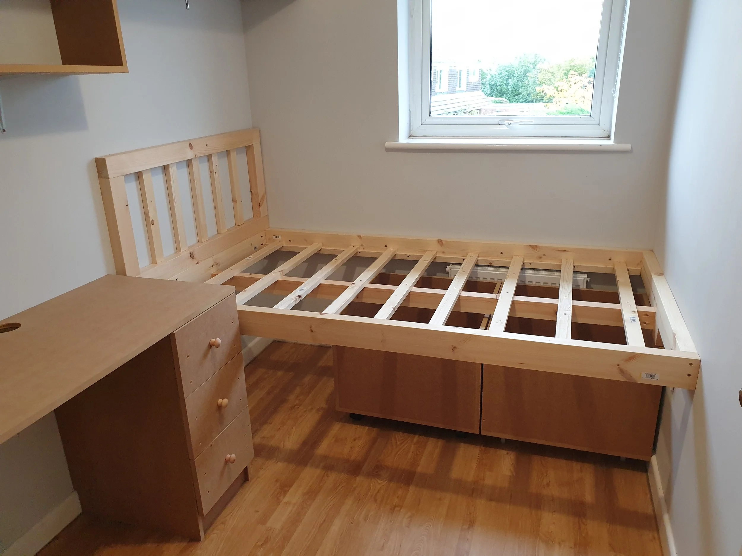 Empty bedroom with wooden bed frame, small nightstand, and a window showing a view of trees outside.