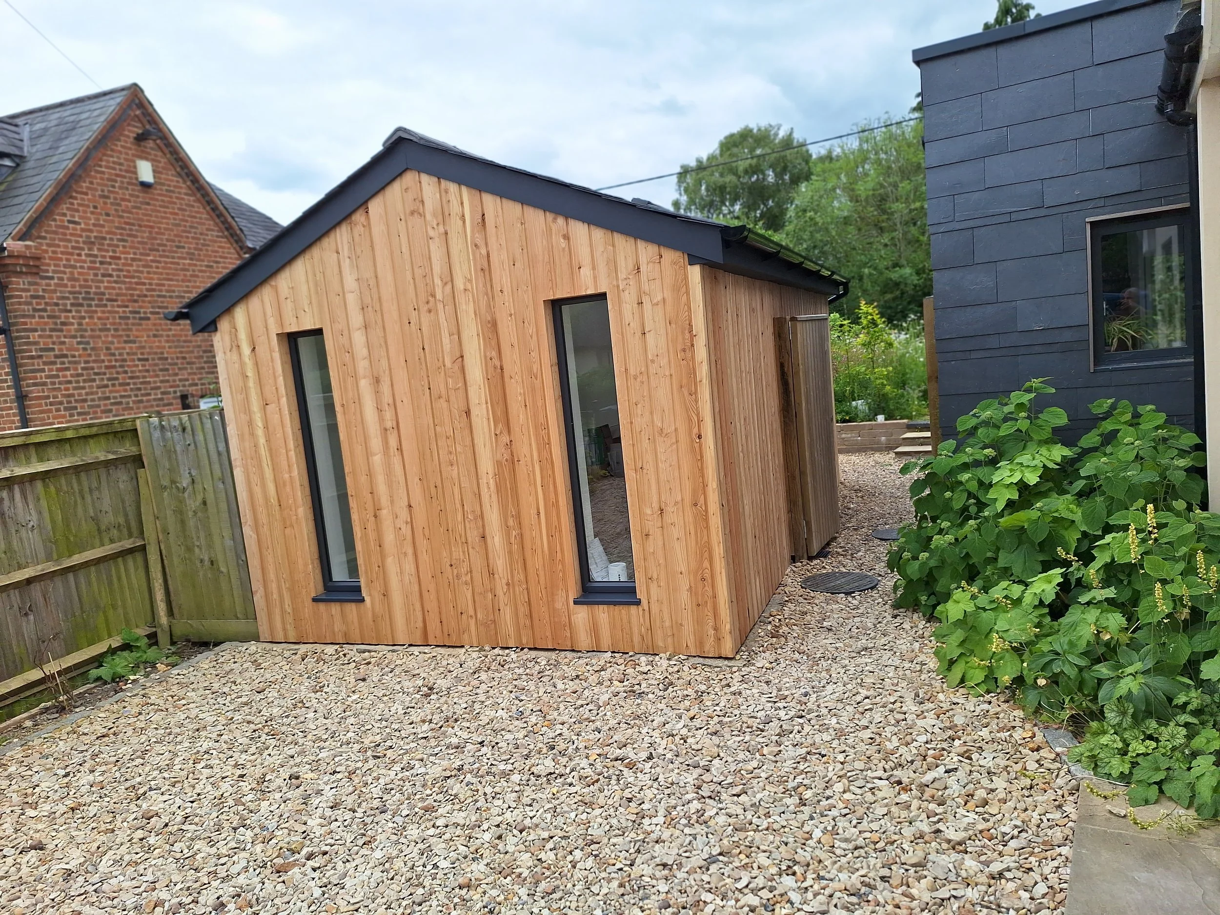 Small wooden shed with vertical planks, black window frames, and a sloped black roof, situated on a gravel yard next to a dark blue house with a window and greenery.