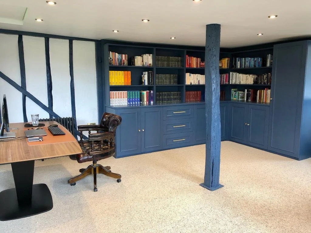 A home office with a wooden desk, computer, and leather office chair. Behind the desk is a built-in dark blue bookshelf filled with books. The room has a beige carpet and white walls with dark wood beams. A blue supporting post is in the middle of th
