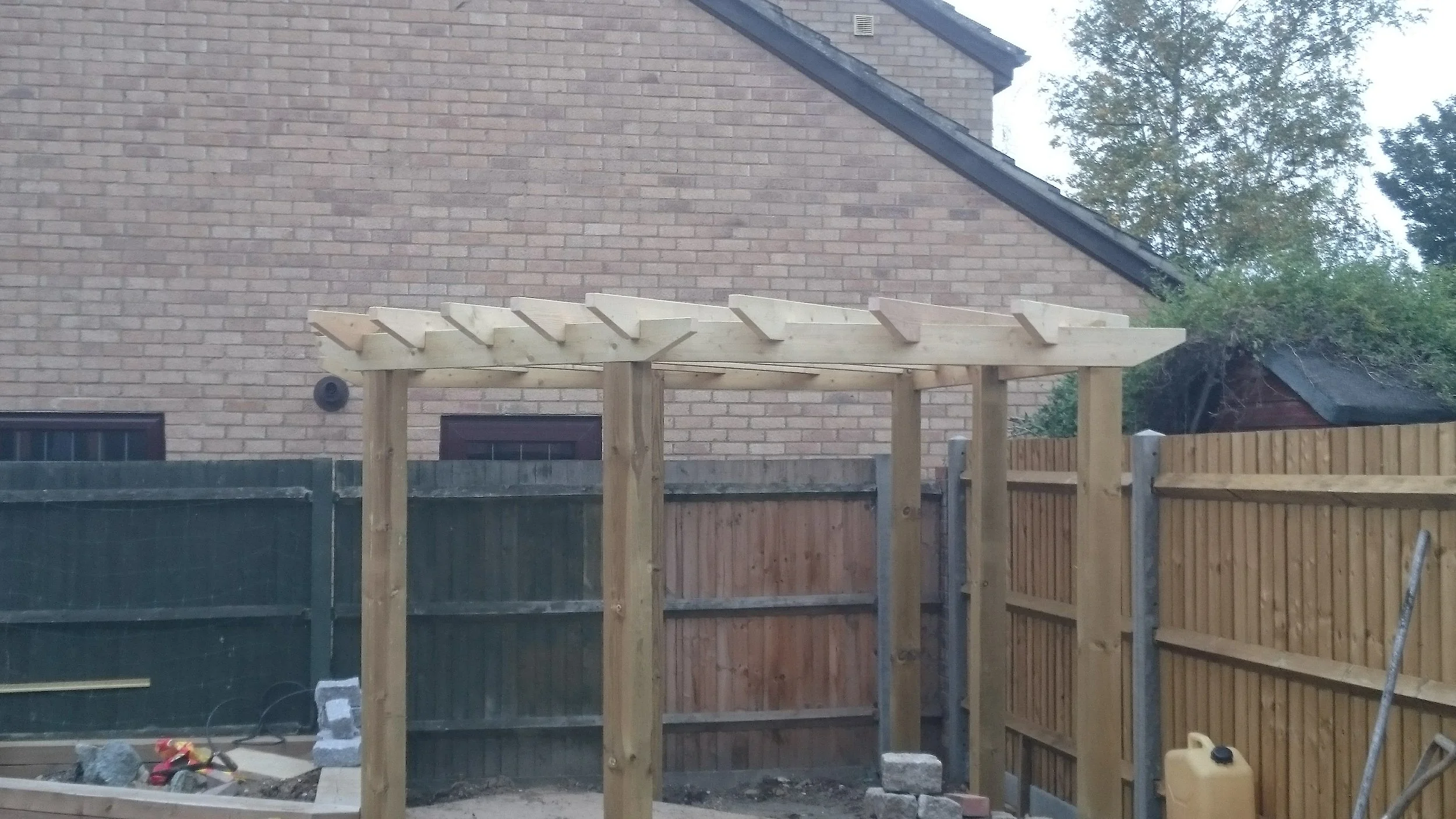 Wooden pergola under construction in a backyard with a brick house, green and brown fence, and trees in the background.