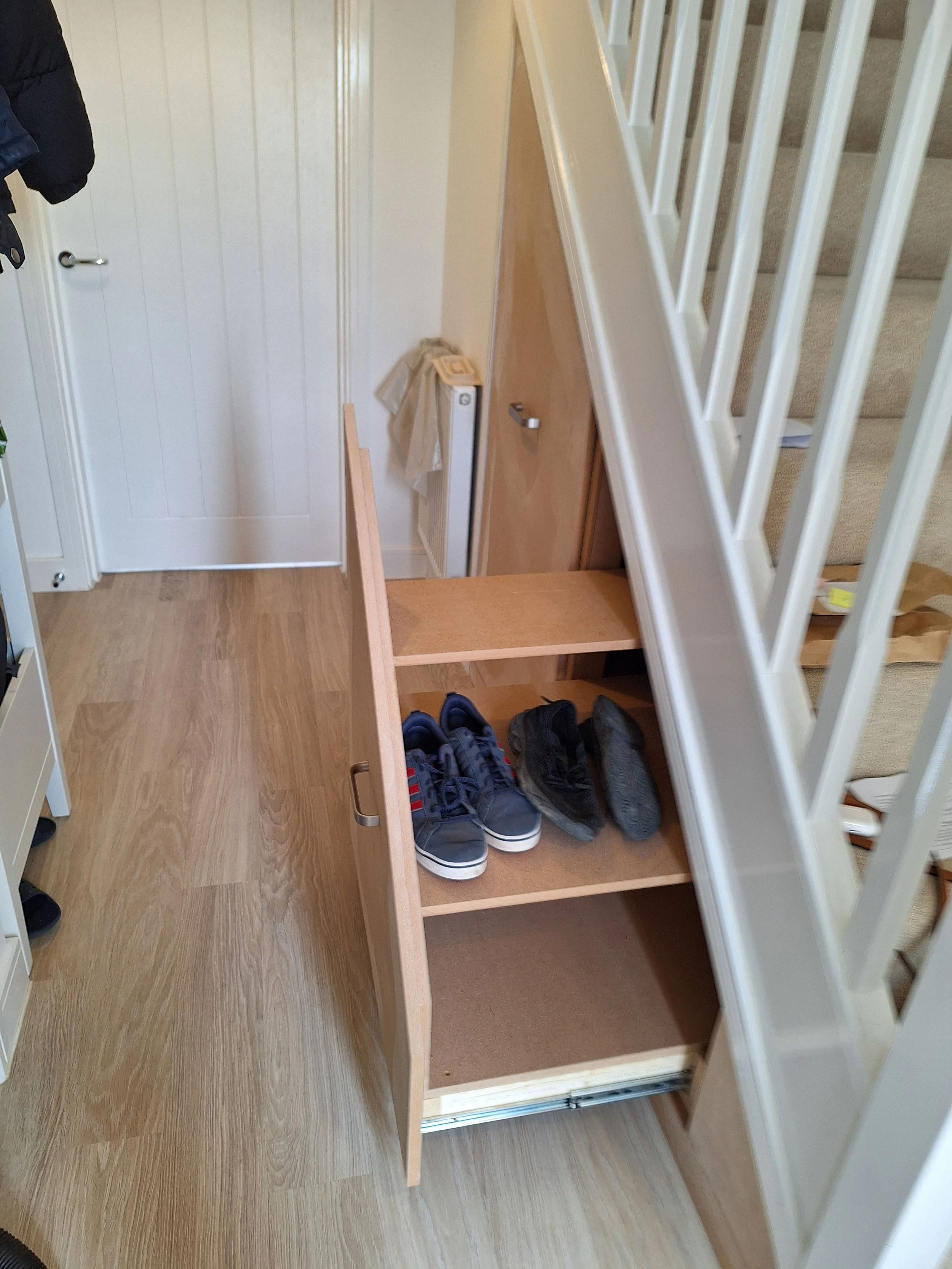 A wooden shoe storage cabinet with two open shelves containing three pairs of sneakers, located at the base of a staircase with white railings.
