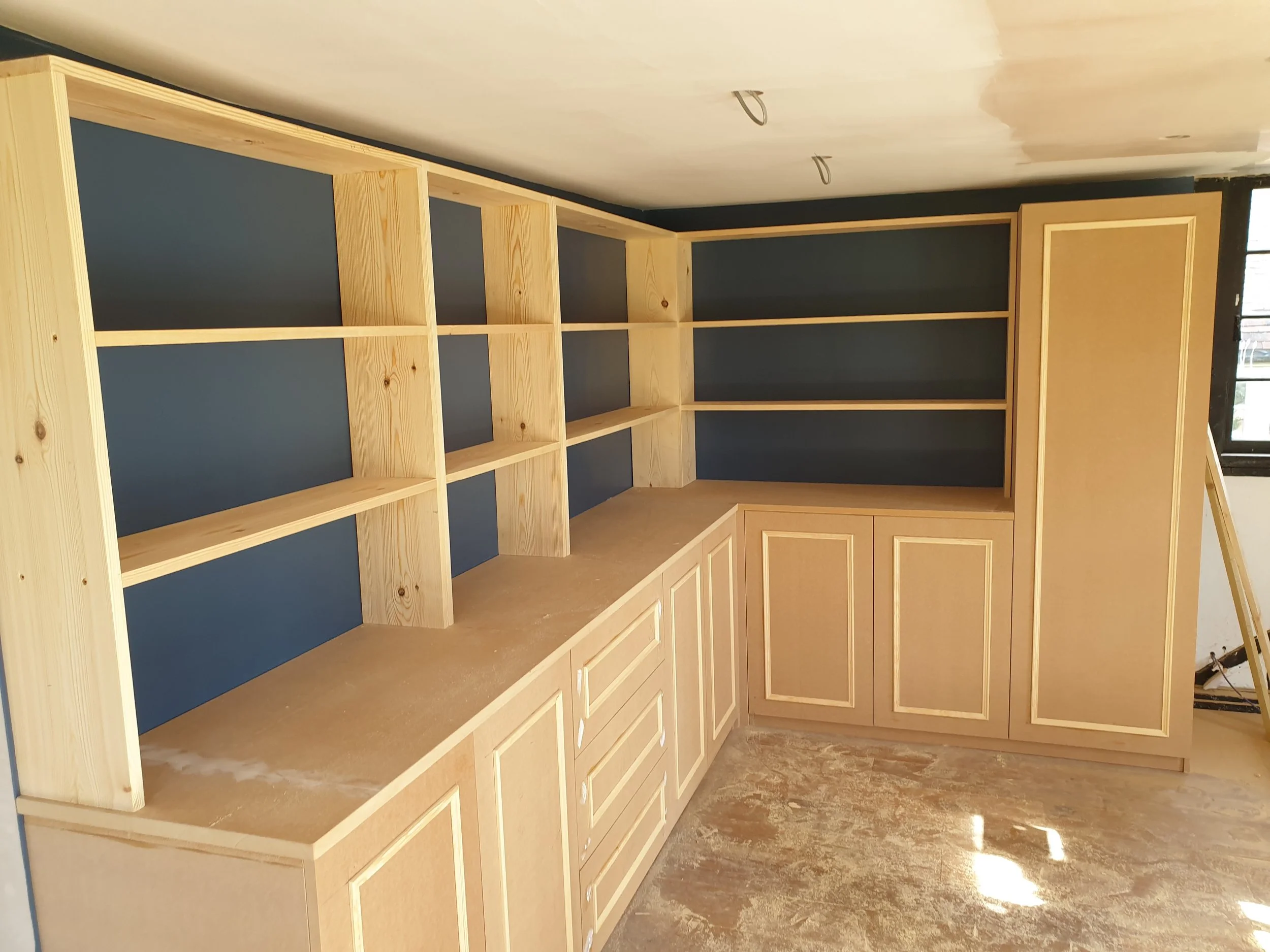 Wooden kitchen or cabinet shelving unit with multiple open and closed storage areas, set against a blue wall with a window, under construction.
