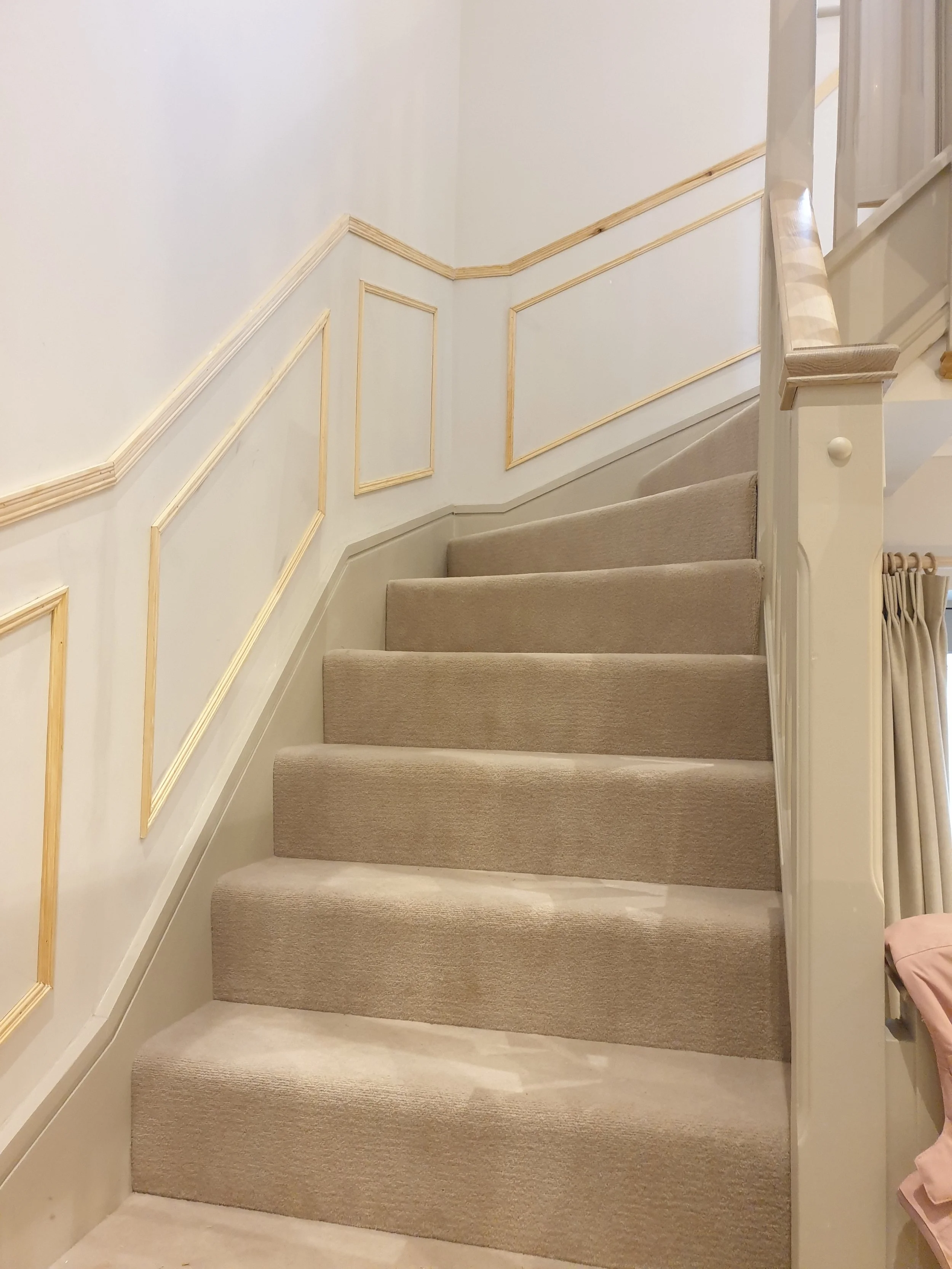 A staircase with beige carpeted steps and a white wooden railing with decorative molding, leading to an upper floor. The wall beside the staircase has white paneling with gold trim details.