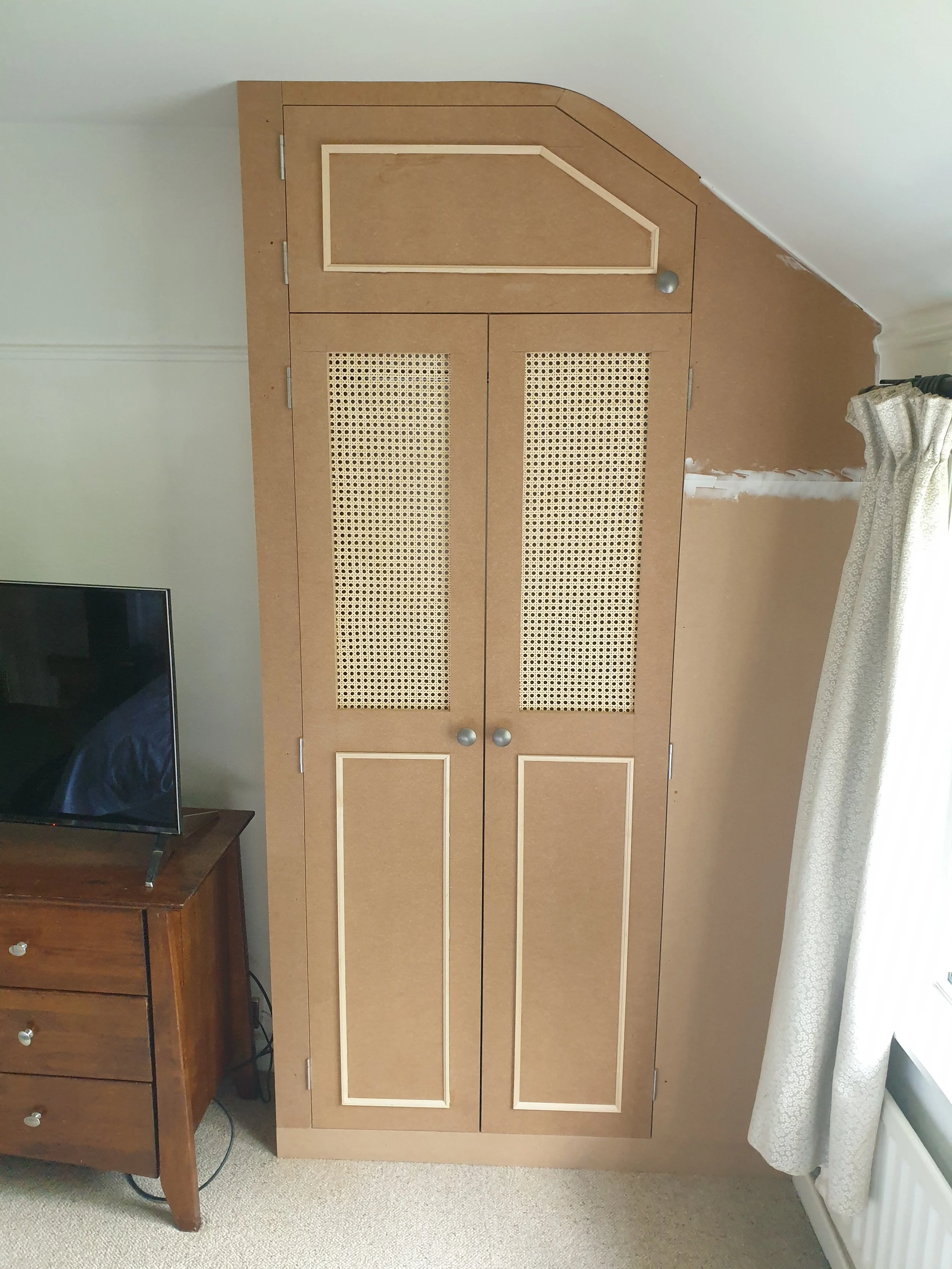 A DIY cabinet with two tall doors with woven cane panels, topped with a smaller door on an angled wall in a room.