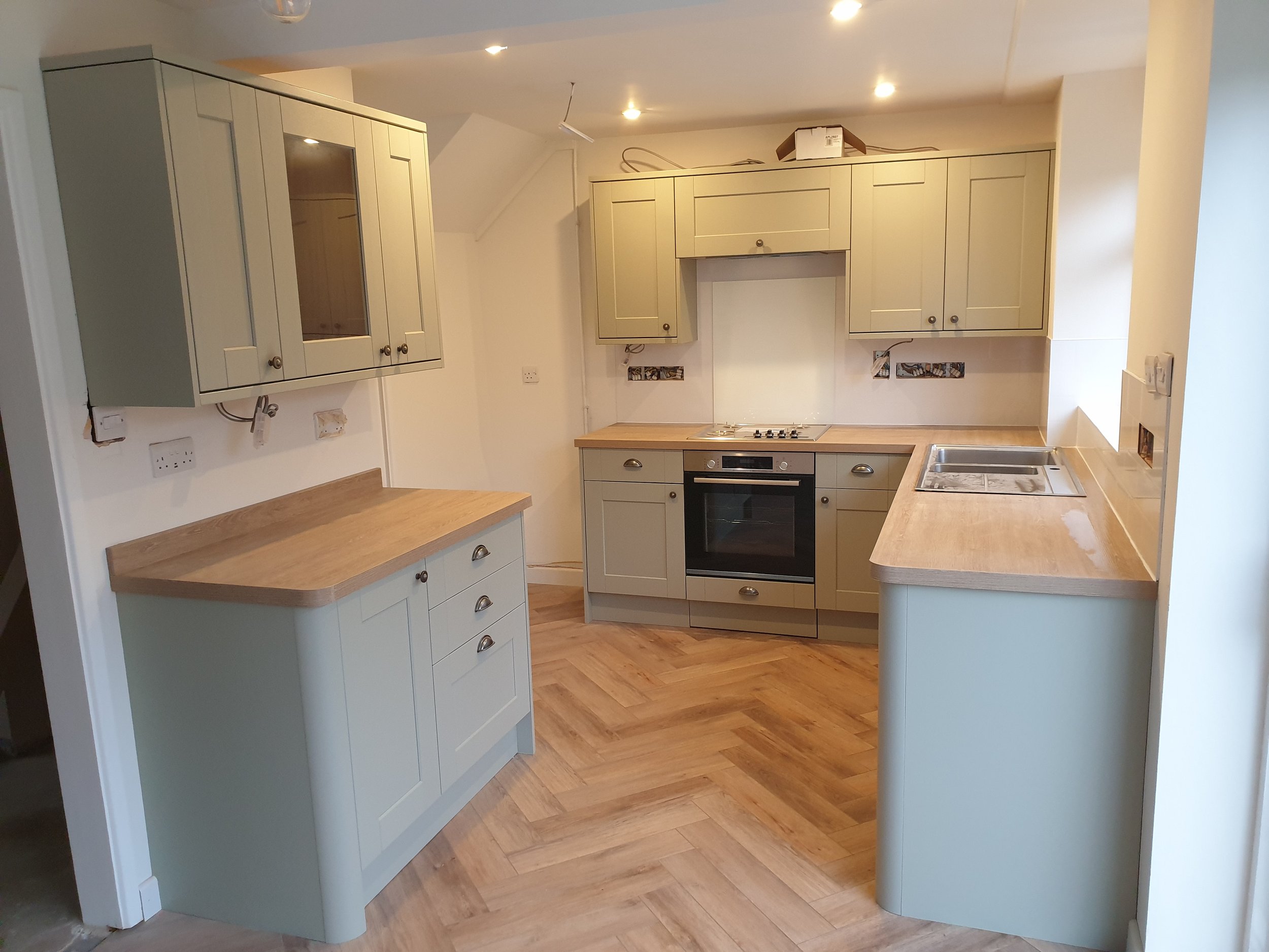 Unfinished kitchen with light-colored cabinets, wooden countertops, and a gas stove with oven. The floor is wood, and there are multiple electrical outlets and wires exposed.