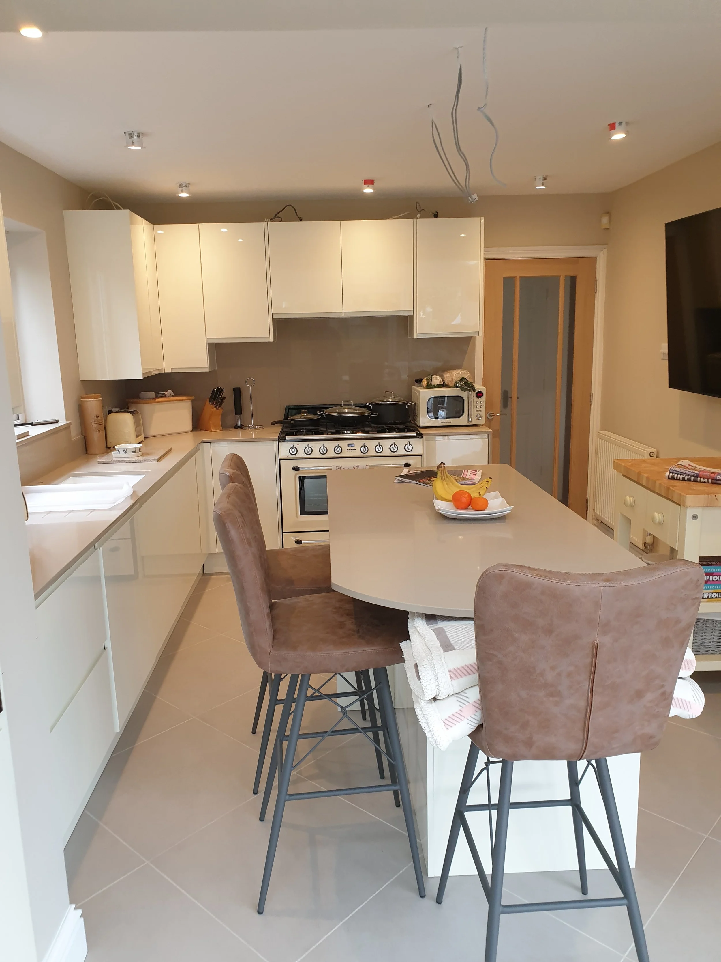A modern kitchen with beige cabinets, a white stove, a microwave, and a countertop with a fruit bowl. There are bar stools around a kitchen island with a towel hanging from it. Ceiling wires are visible, indicating ongoing installation or repairs.