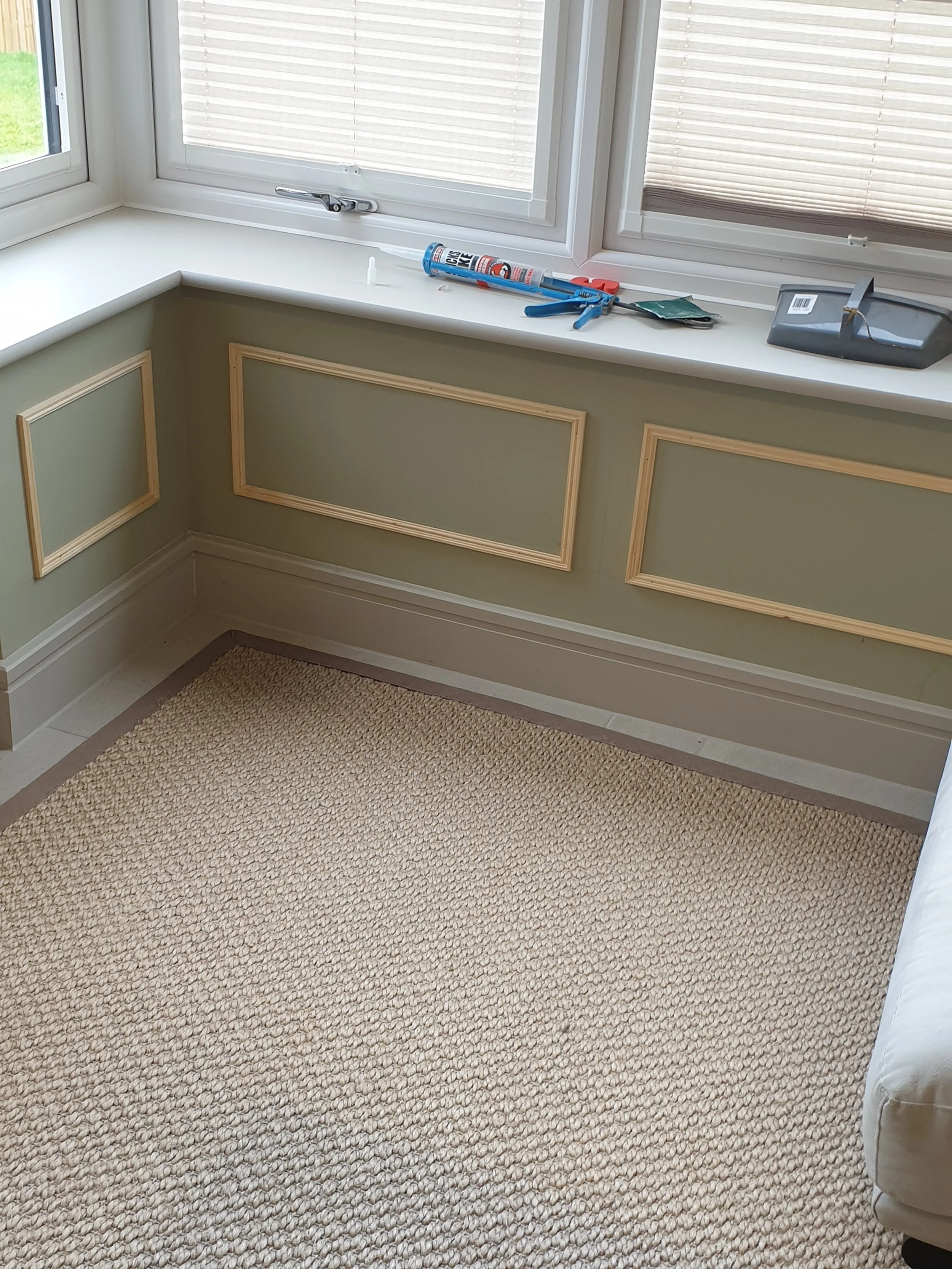 Window sill with tools and materials for home repair, including caulk, a utility knife, and glue, near green paneled walls and a beige carpeted floor.