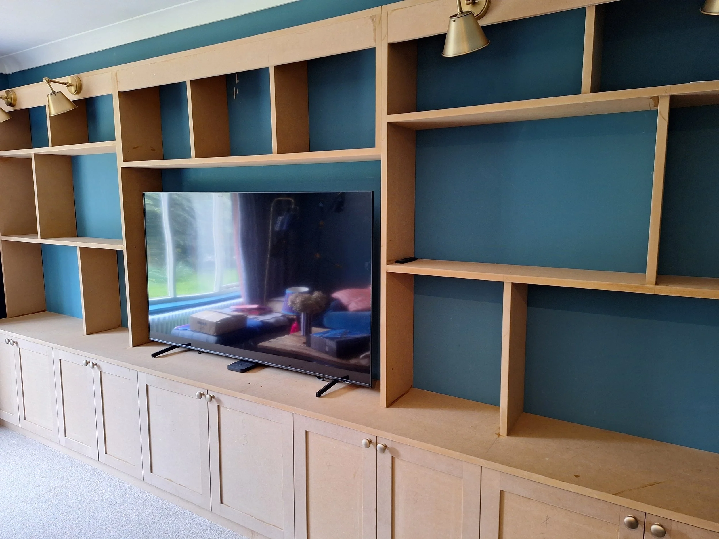 Empty wooden shelving unit with blue backing and a flat-screen TV on the bottom left shelf, reflecting a window and some objects in the room.