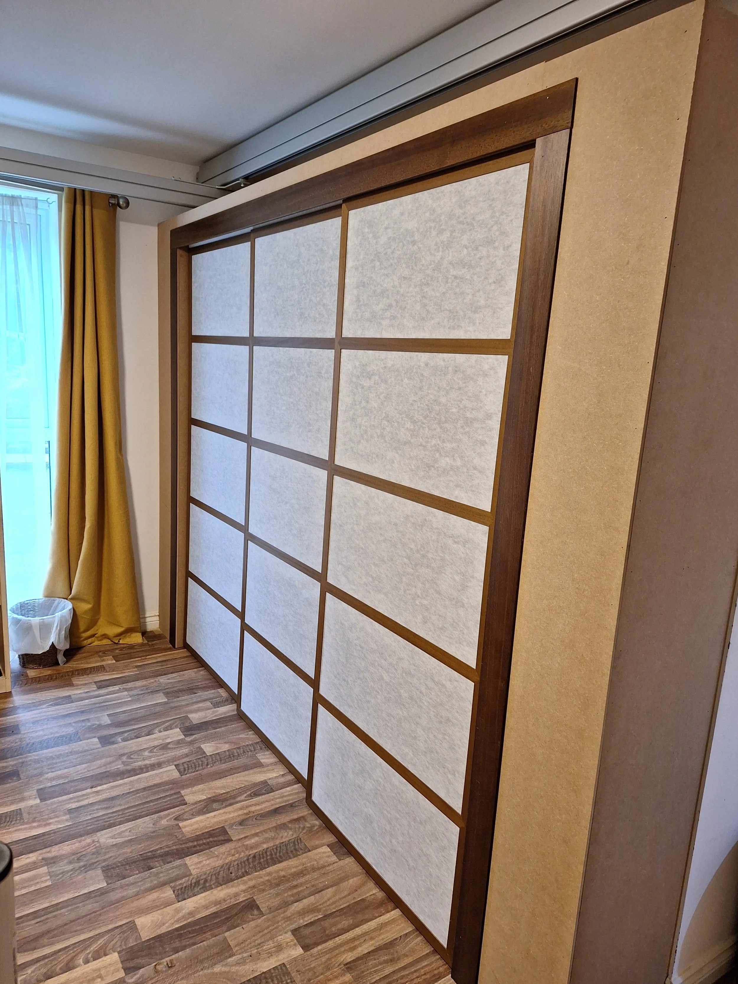 A large wooden and paper sliding door, typical of traditional Japanese shoji, situated in a room with wooden flooring and yellow curtains near a window.