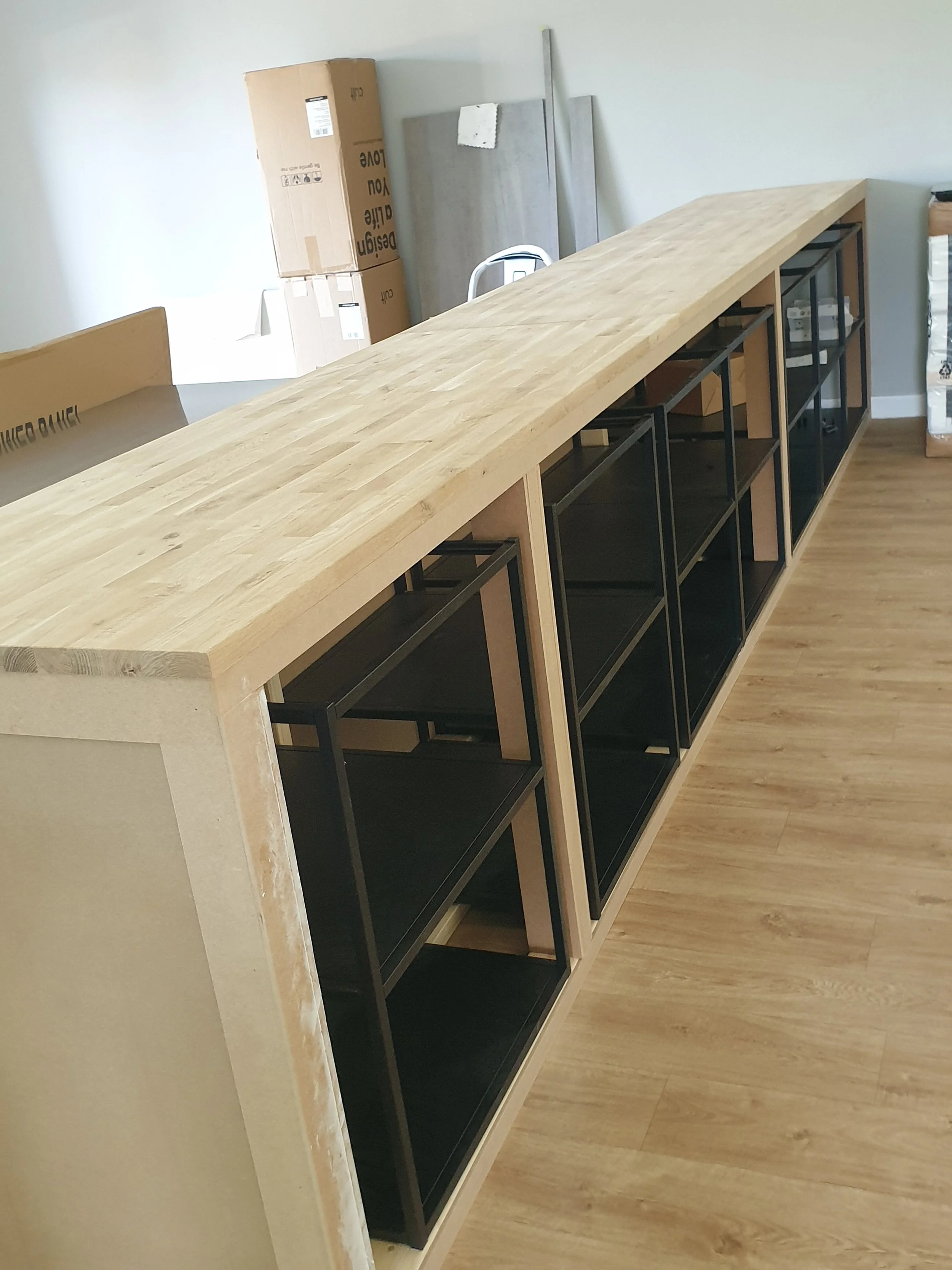 A partially assembled wooden and black metal shelving unit with a wooden countertop on top, in a room with light-colored walls and wood flooring. Cardboard boxes and construction materials are visible in the background.