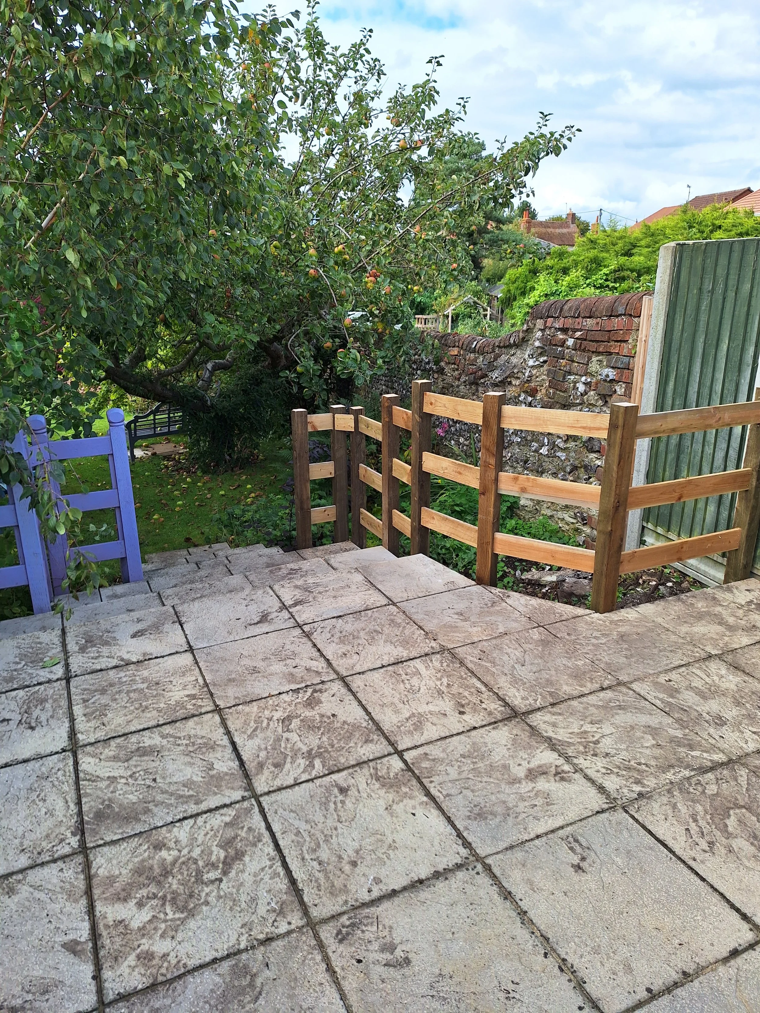 A backyard patio with beige stone tiles, surrounded by a wooden fence and greenery, including a tree with apples, a brick wall, and a green shed.