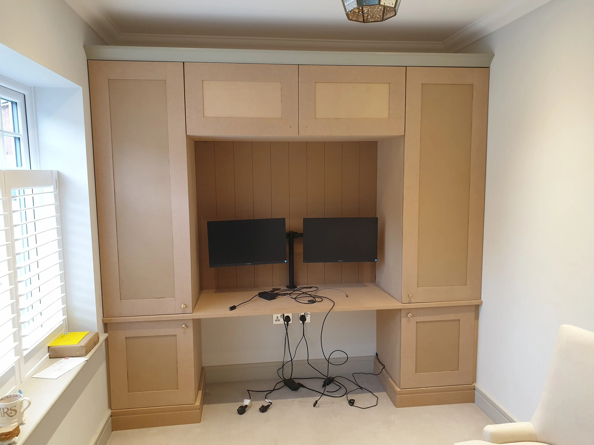 A built-in wooden wall unit with cabinets, a shelf, and a workspace with two monitors, laptop, and tangled cables in a room with a window and white walls.