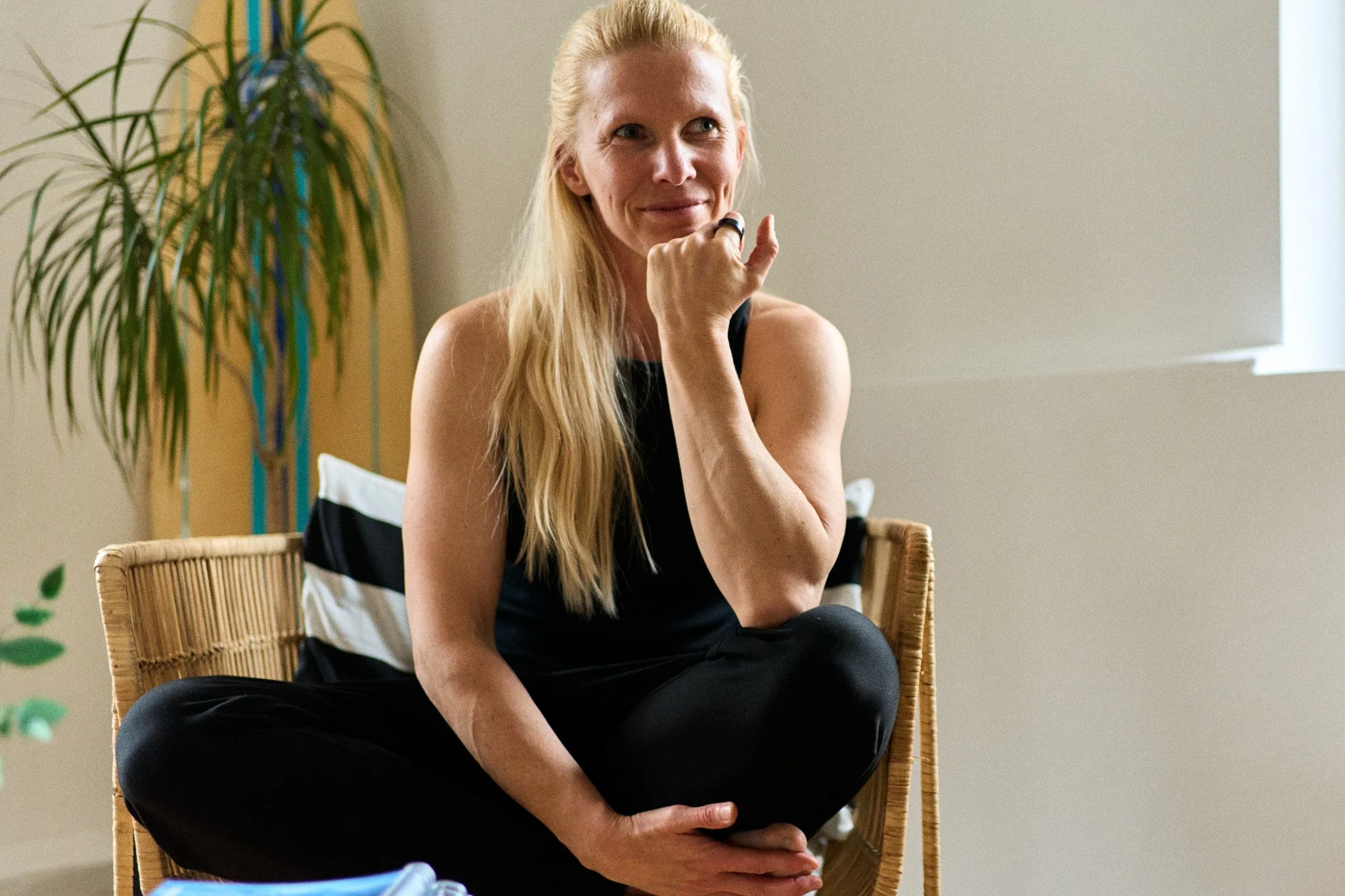 Eine blonde Frau sitzt lächelnd auf einem Stuhl in einem Raum mit neutralen Wänden, neben einer Pflanze, die im Hintergrund steht. Sie trägt ein schwarzes Top und schwarze Hosen und wirkt entspannt.