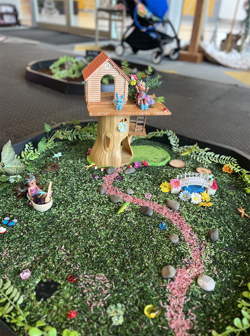 Miniature fairy garden with a treehouse, fairy figurines, a small pond with a bridge, and decorative plants, set on a table in a store or outdoor setting.
