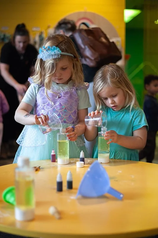 Two young girls wearing colorful costumes, possibly for a themed event, are conducting a science experiment with liquids at a table in a lively indoor setting.