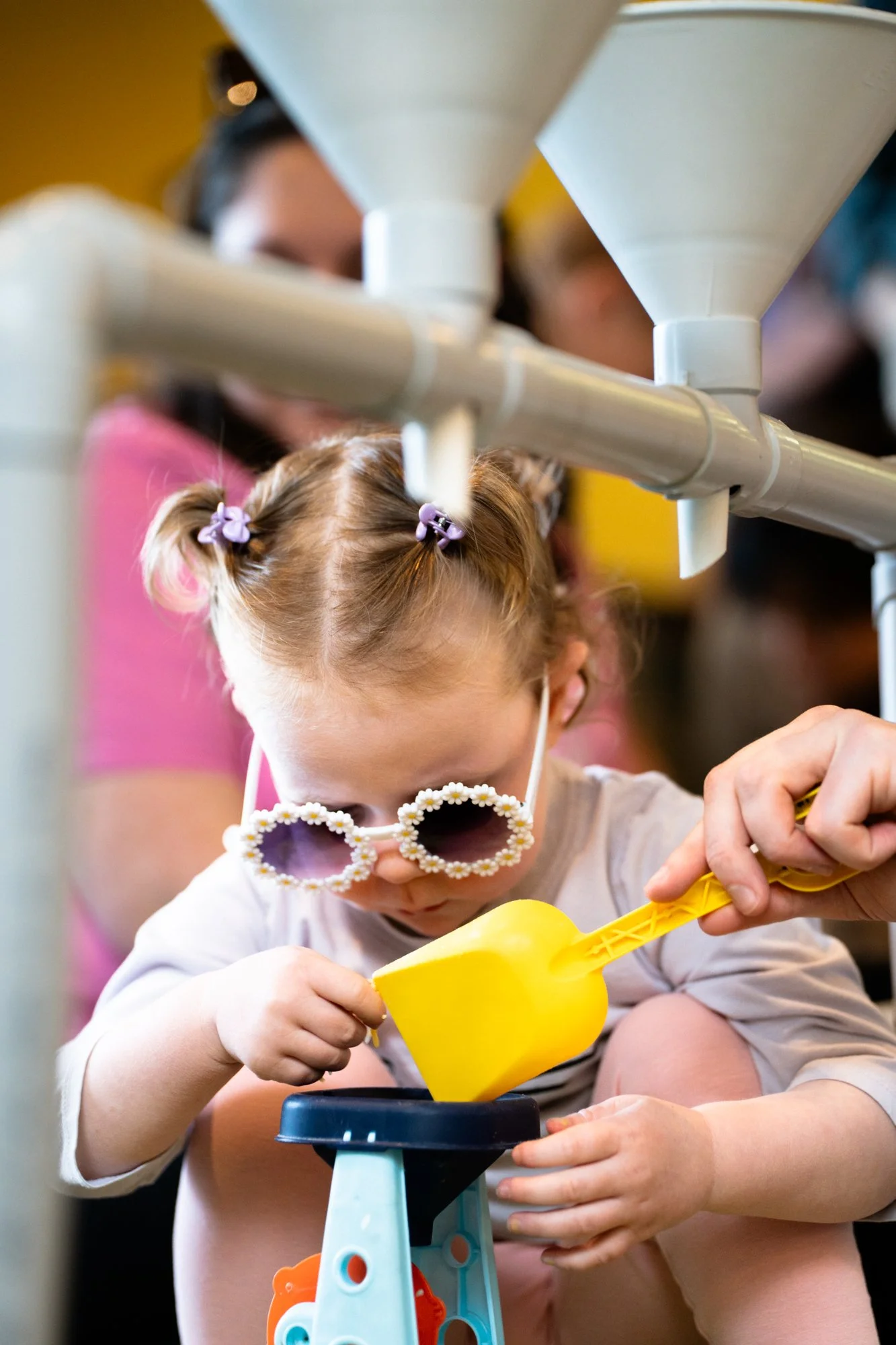 A young girl with blonde hair in pigtails and sunglasses shaped like flowers, playing with a yellow toy shovel, with adults in the background.
