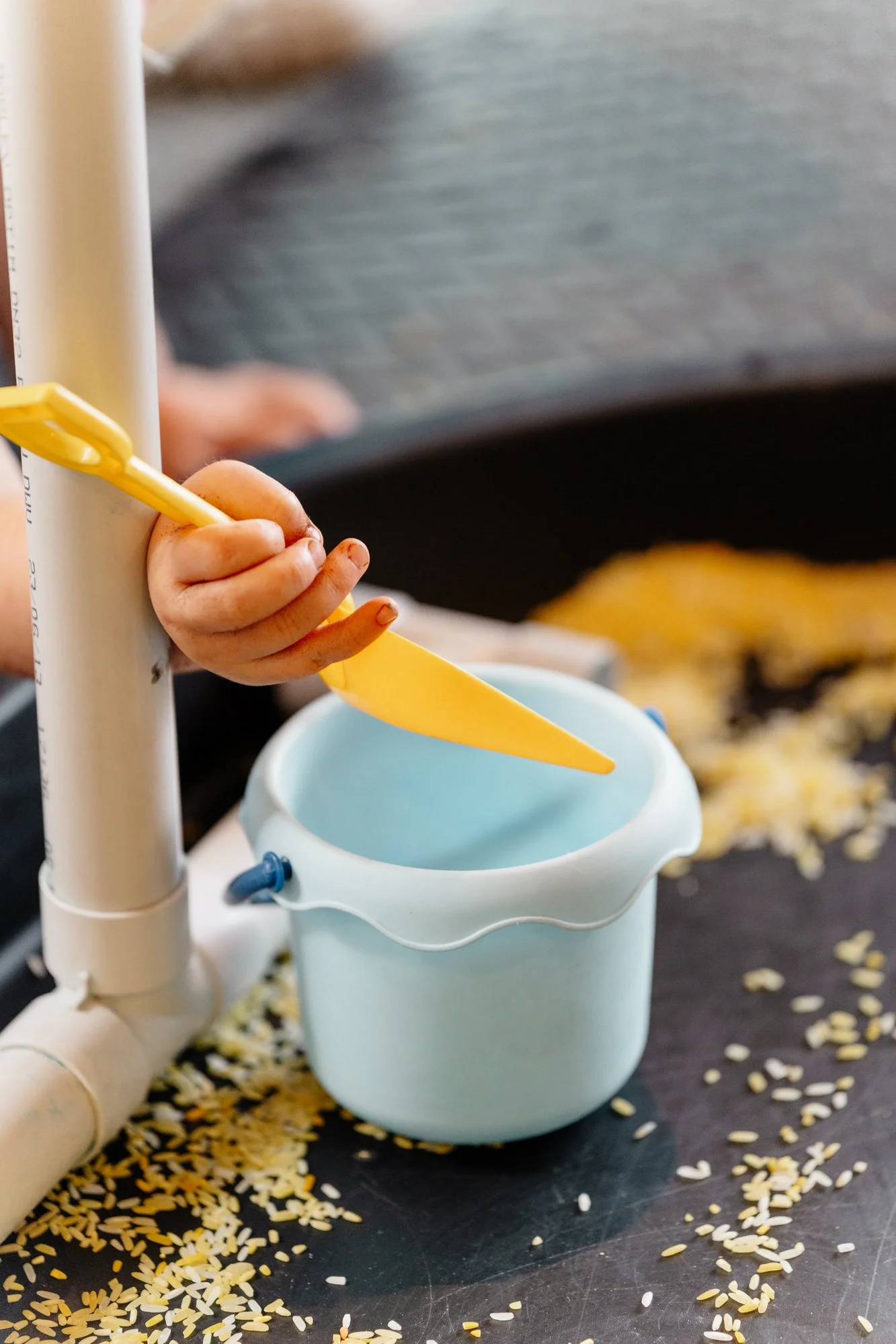 A child's hand holds a yellow spatula, stirring rice in a small white bowl. Rice is scattered around on a dark surface in the background.