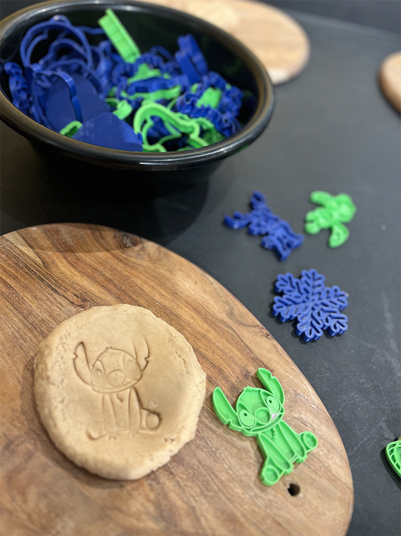 A wooden cutting board with a Christmas-type cookie featuring an reindeer etched into the top. Next to the cookie is a green cookie cutter shaped like a reindeer. In the background, a black bowl is filled with blue and green snowflake-shaped and reindeer-shaped cookie cutters, some on the dark surface surrounding the cutting board.